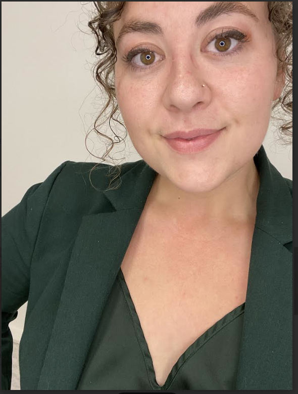 Close-up of a woman with curly hair and a nose piercing, wearing a dark green blazer and top, smiling at the camera.