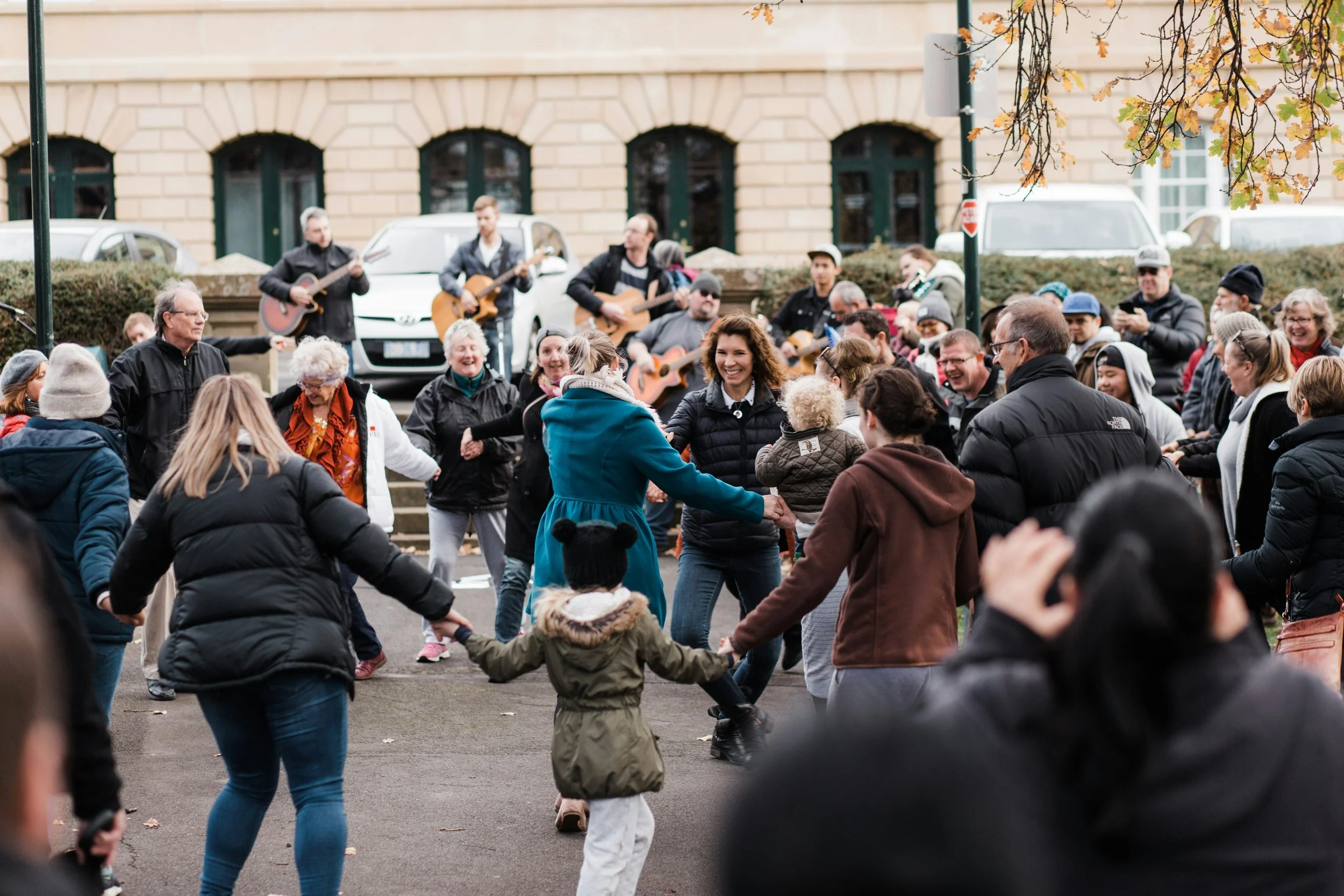 People dancing in a circle outdoors, with a band playing guitars in the background, during a festive gathering.