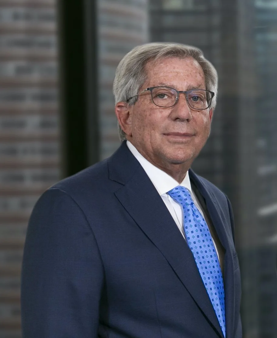 An older man with gray hair and glasses wearing a dark blue suit, white shirt, and light blue tie, standing in front of a blurred cityscape.