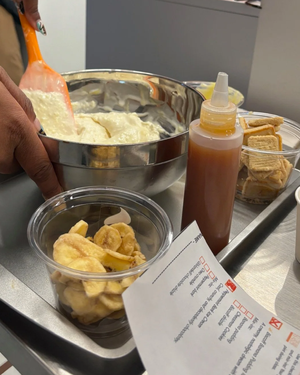Preparation of fried plantains with a bowl of mashed potatoes, a bottle of sauce, and a container of toasted sandwiches on a metal tray.