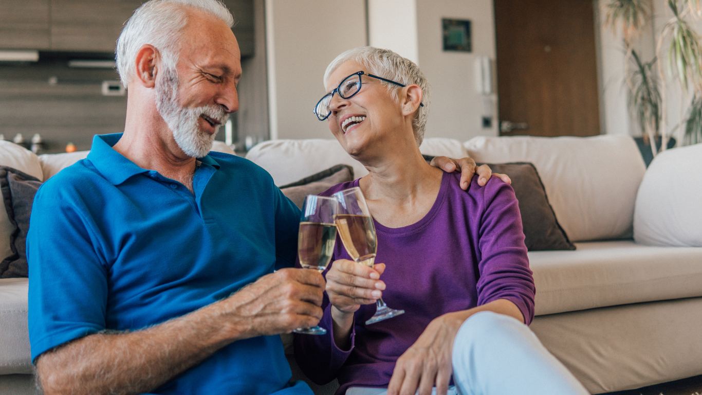 An elderly couple sitting on the floor in front of a sofa, smiling and laughing as they toast glasses of champagne.