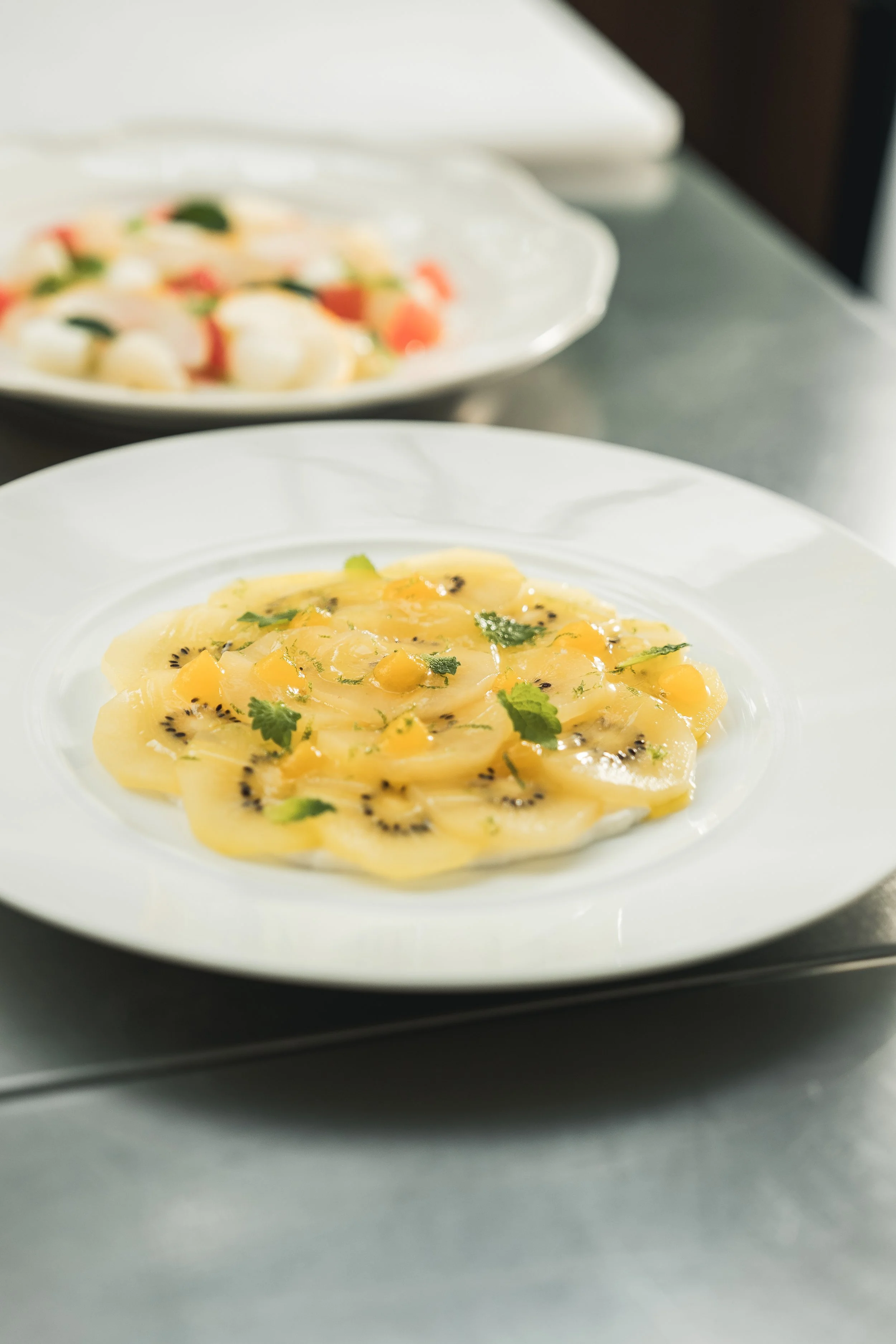 A plate of sliced yellow fruit, possibly peaches or mango, garnished with black sesame seeds and cilantro. In the background, a blurry dish with white balls and assorted vegetables is visible.