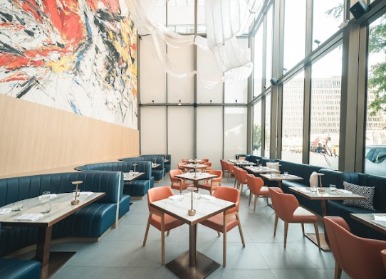 High end sophisticated restaurant with artistic setting and large floor to ceiling windows with marble tables and city view, colorful abstract fresco art on wall.
