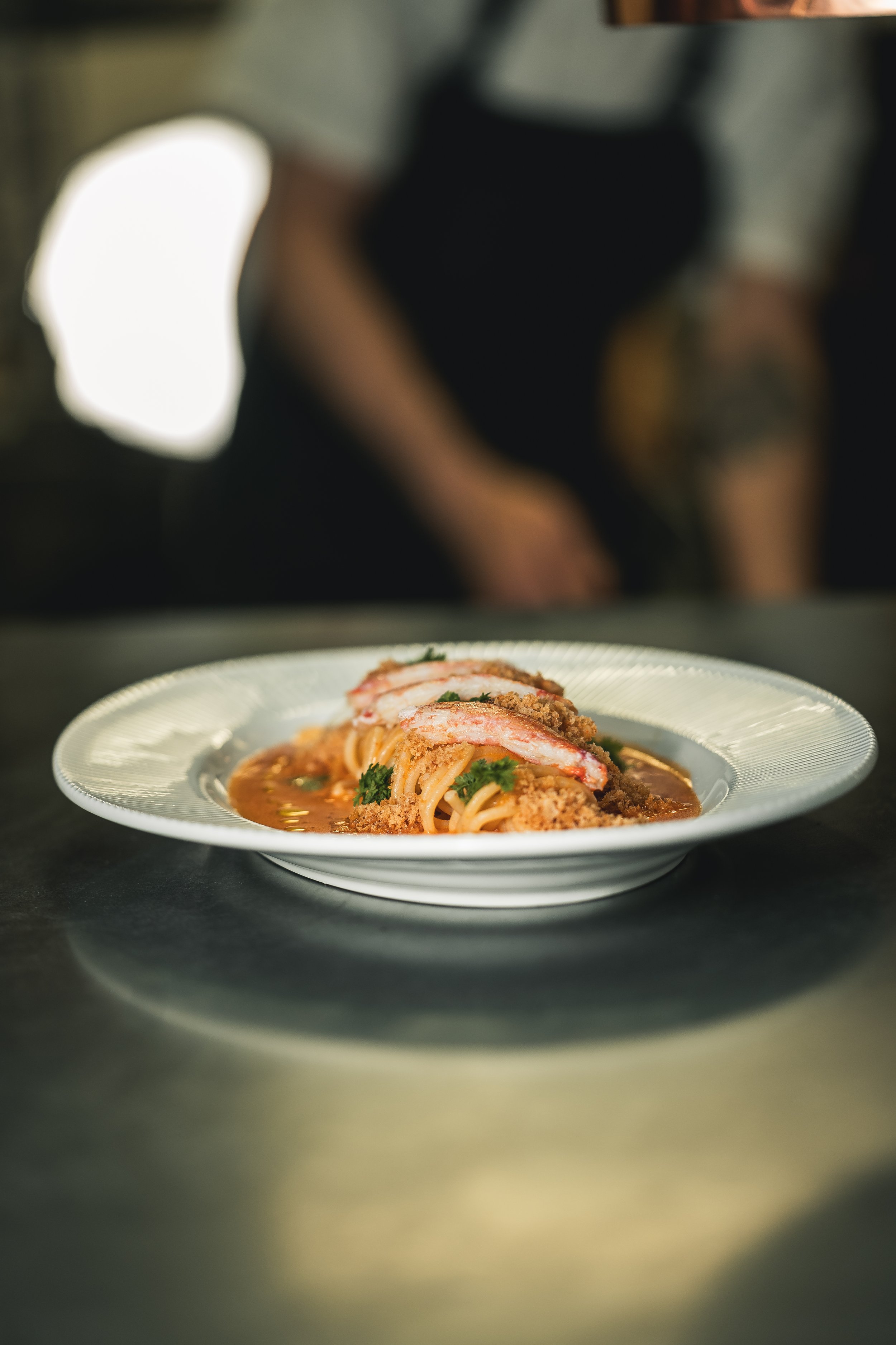 Close-up of a plate of pasta with breadcrumb topping, garnished with parsley, with a blurred background including a person in a black shirt.