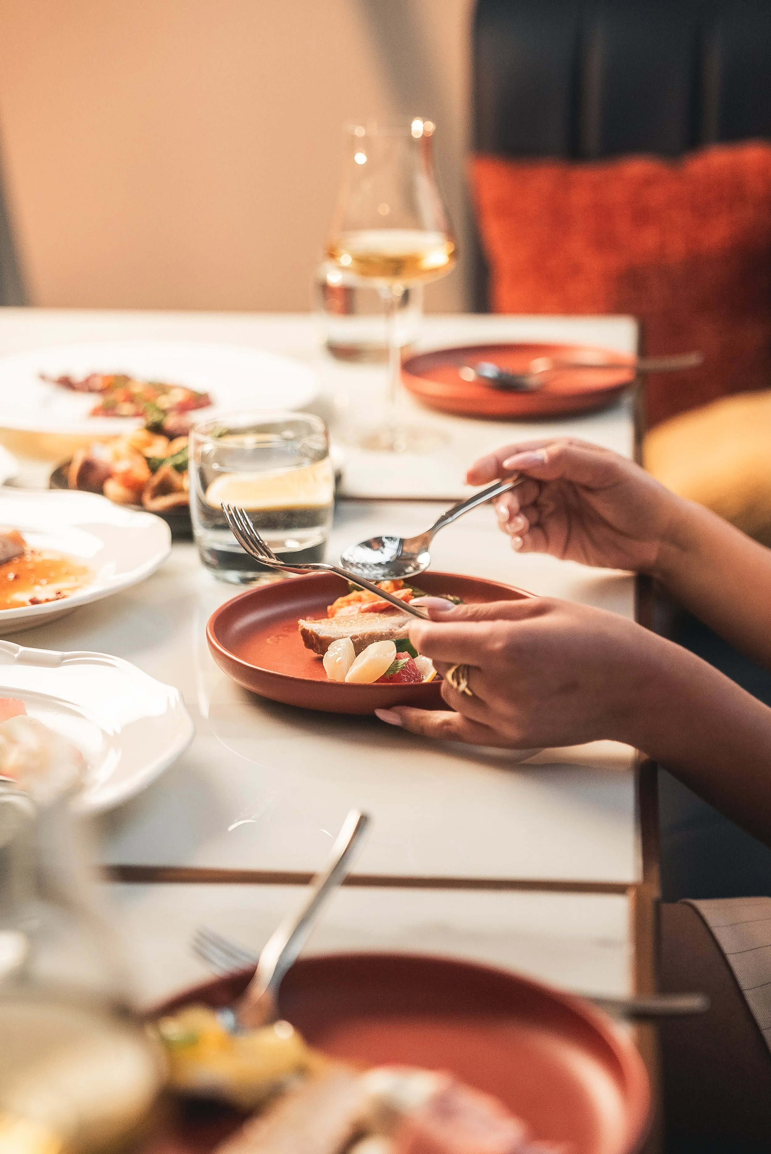 A person holding a spoon over a bowl of food, with a dinner table set with various dishes, a glass of white wine, and a water glass in the background.