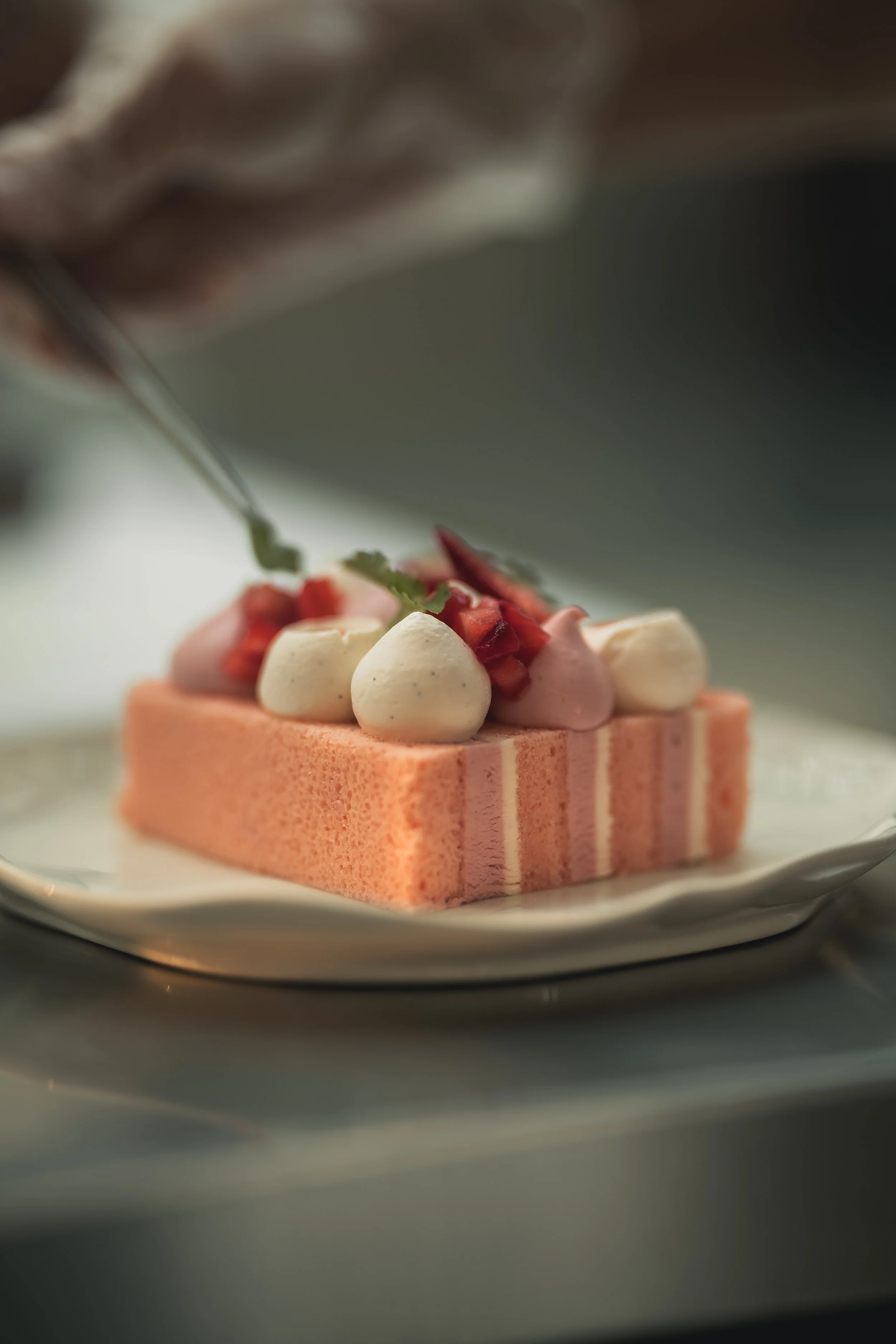 Close-up of a pink and white layered dessert with small dollops of cream and chopped strawberries, garnished with a mint leaf, on a white plate.