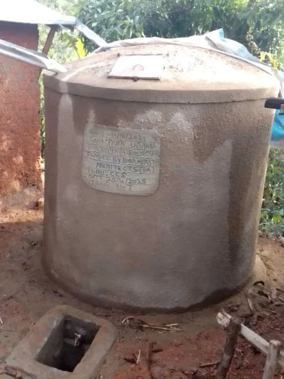 A large, round, concrete water tank with a lid and a handwritten label attached to its surface, set outdoors on dirt ground surrounded by plants and a partial view of a building.