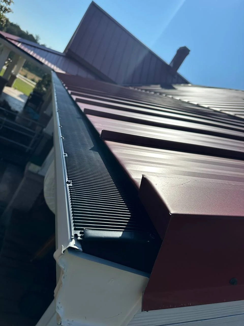 Close-up of the edge of a metal roof with vertical brown panels and an air conditioning unit with a white exterior and black grille over a sunny outdoor background.