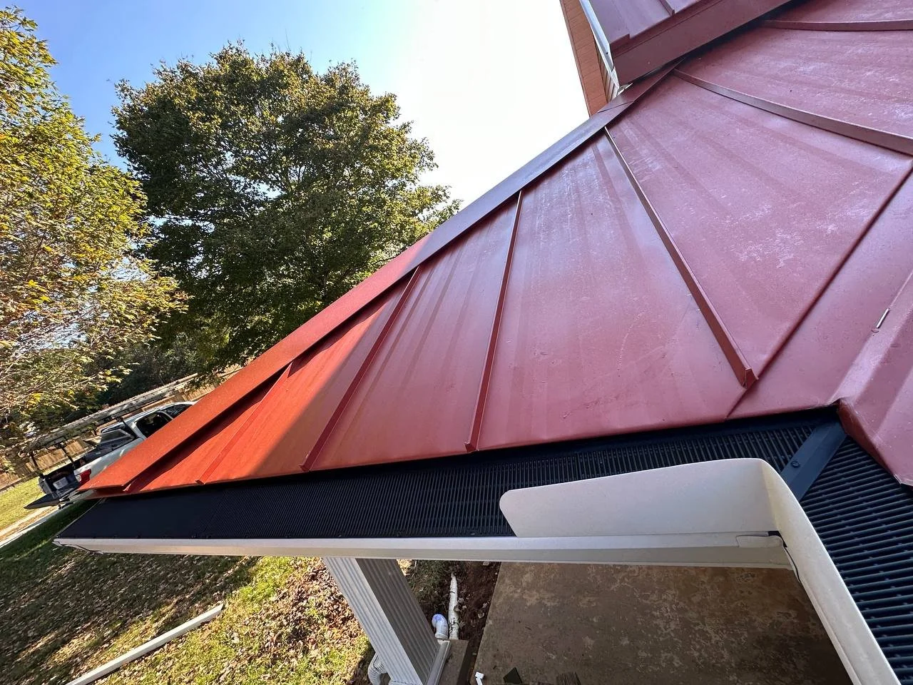 Close-up of a red metal roof with a black trim, part of a house, with trees and a blue sky in the background.