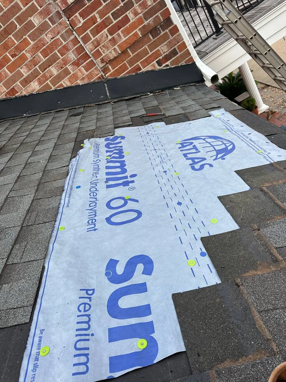 Installing synthetic underlayment on a roof with asphalt shingles, with a brick chimney and a white downspout in the background.