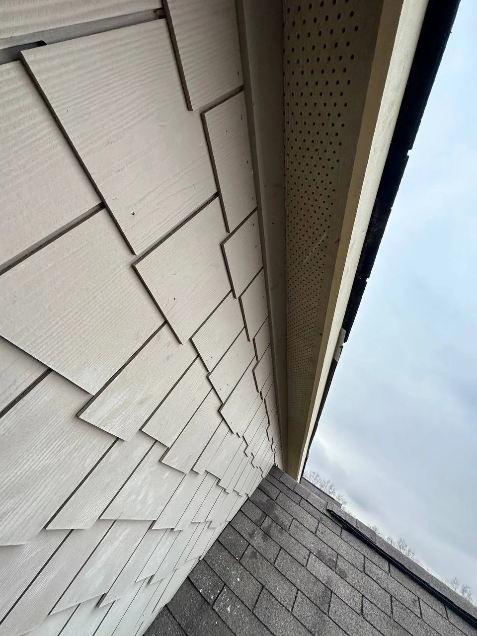 Close-up view of the exterior wall of a house showing beige siding shingles, vented soffit, and part of a shingled roof.