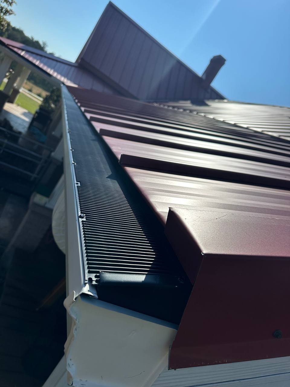Close-up of a metal roof with a vent or ridge cap, reflecting sunlight, with part of a house, backyard, and blue sky in the background.