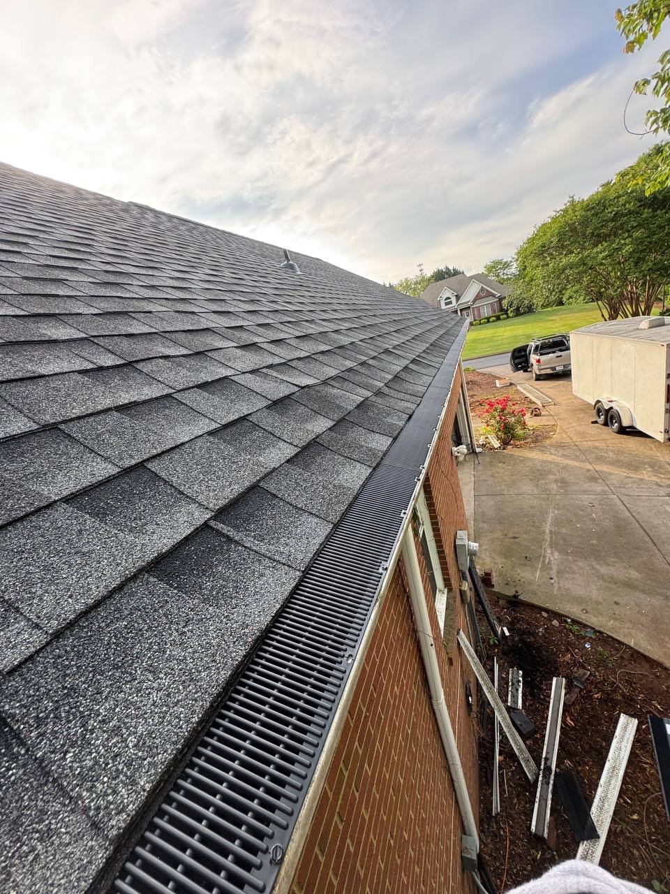 Close-up view of a residential roof with gray shingles and a gutter system, overlooking a backyard with a concrete driveway, a trailer, and a black pickup truck, with trees and house in the background.