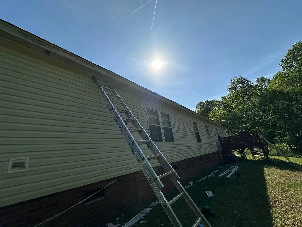 A ladder leaning against the side of a house next to a wooden deck with trees in the background and the sun shining brightly in the sky.