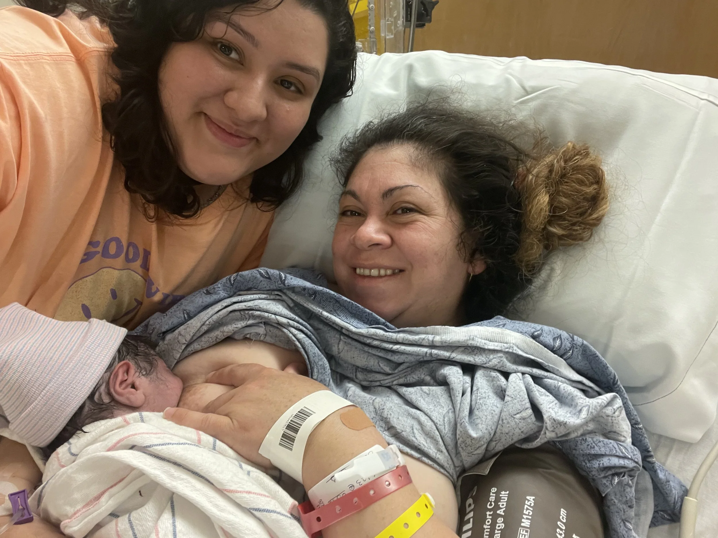 Woman in hospital bed holding newborn baby, with another woman smiling beside her.