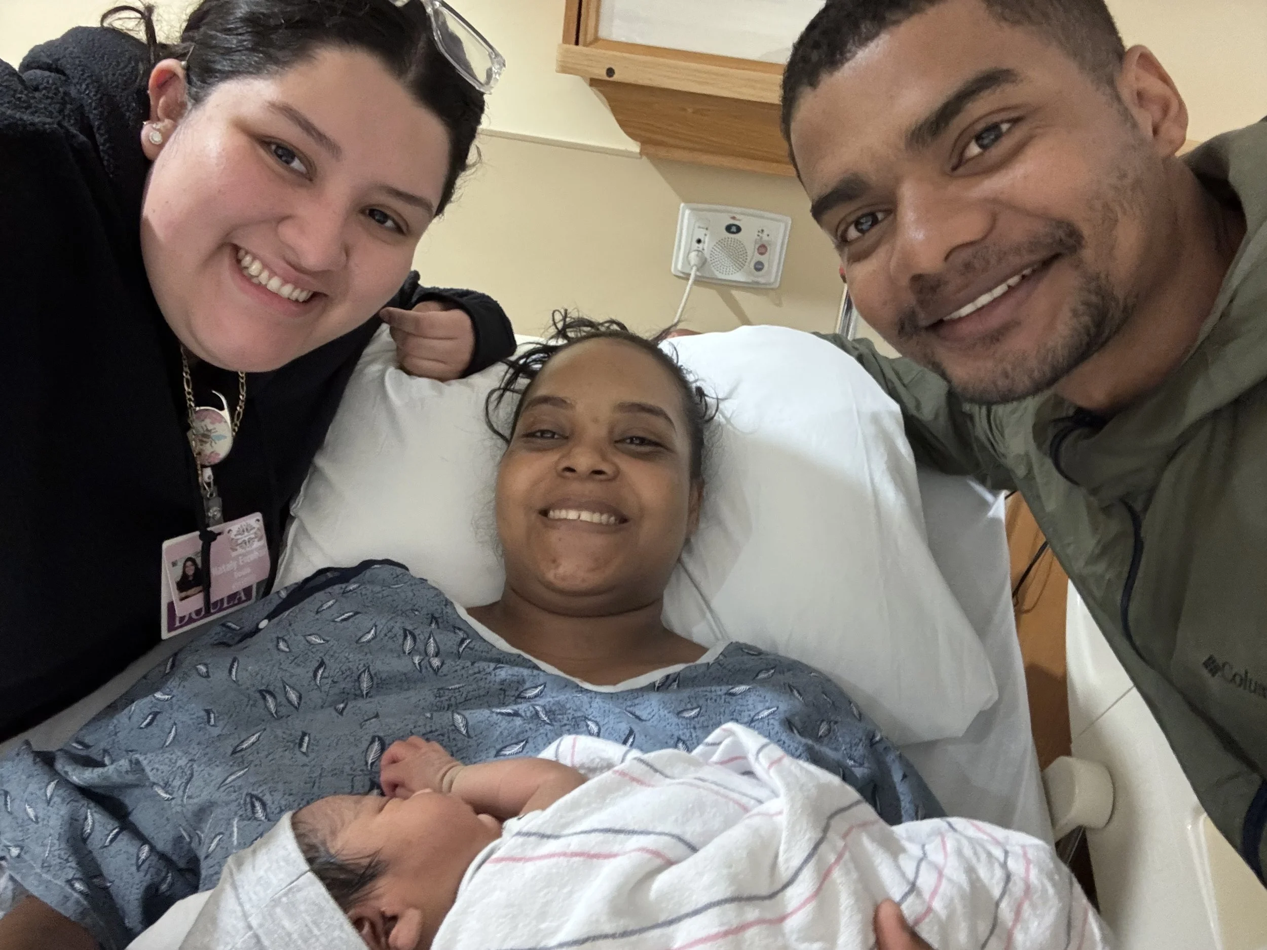 A woman lying in a hospital bed holding a newborn baby, with two visitors smiling on either side of her.
