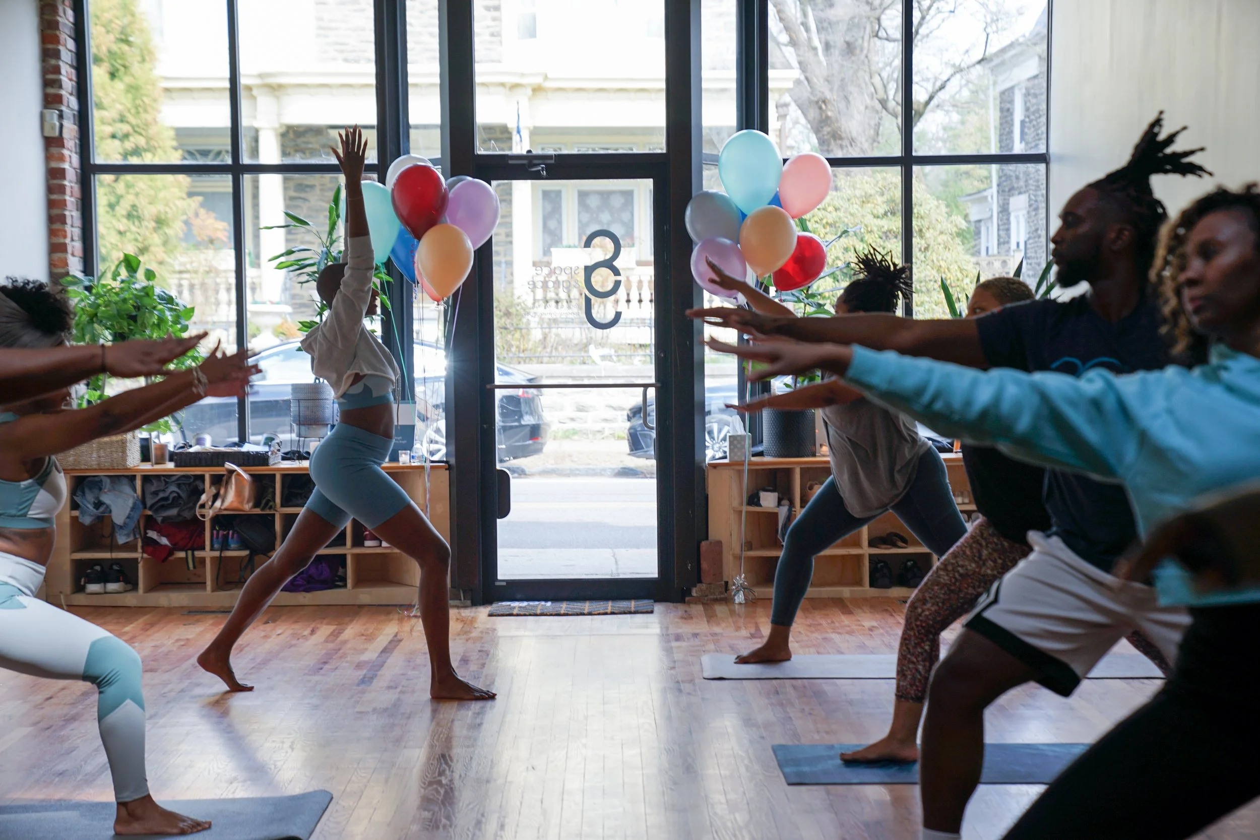 A group of people practicing yoga in a studio with large glass windows, balloons tied to the door, and plants inside.