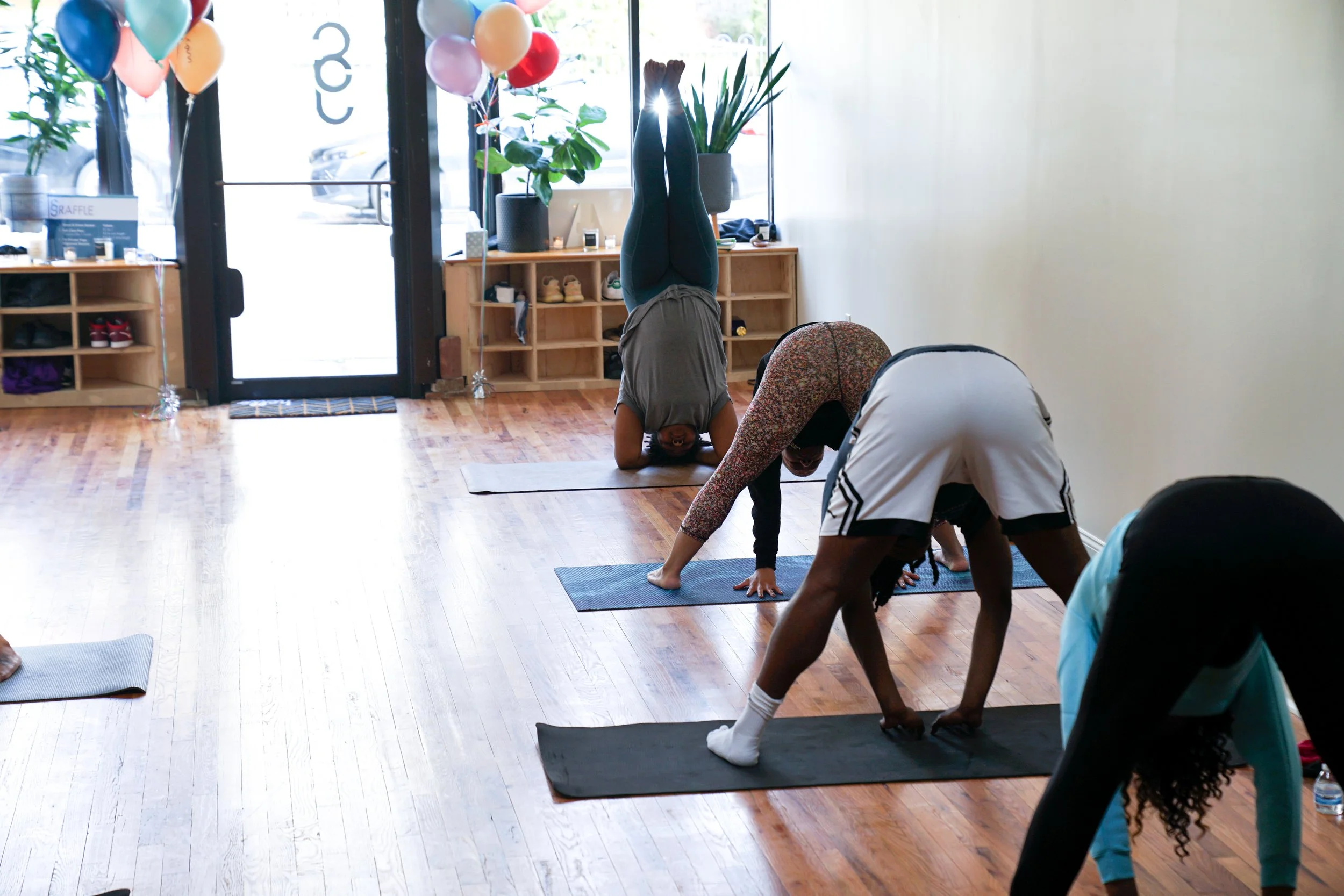 People practicing yoga in a studio with wooden floors, balloons near the entrance, and sunlight coming through the windows.