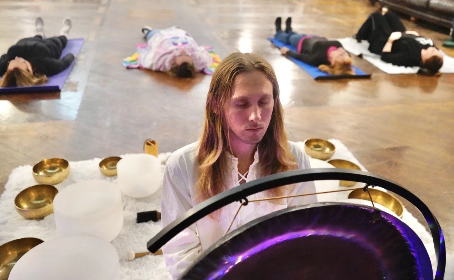 A woman with closed eyes sitting in front of a large singing bowl with purple lighting, surrounded by smaller gold bowls on a white carpet. In the background, four people lie on yoga mats in a dimly lit room with wooden floors.