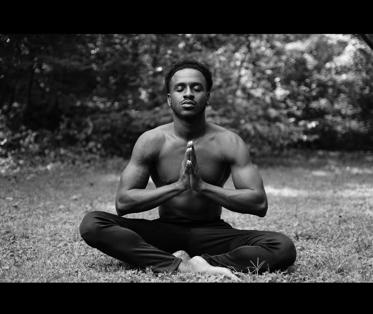 A shirtless young man practicing meditation outdoors, sitting cross-legged on grass with hands in prayer position, eyes closed, surrounded by trees.