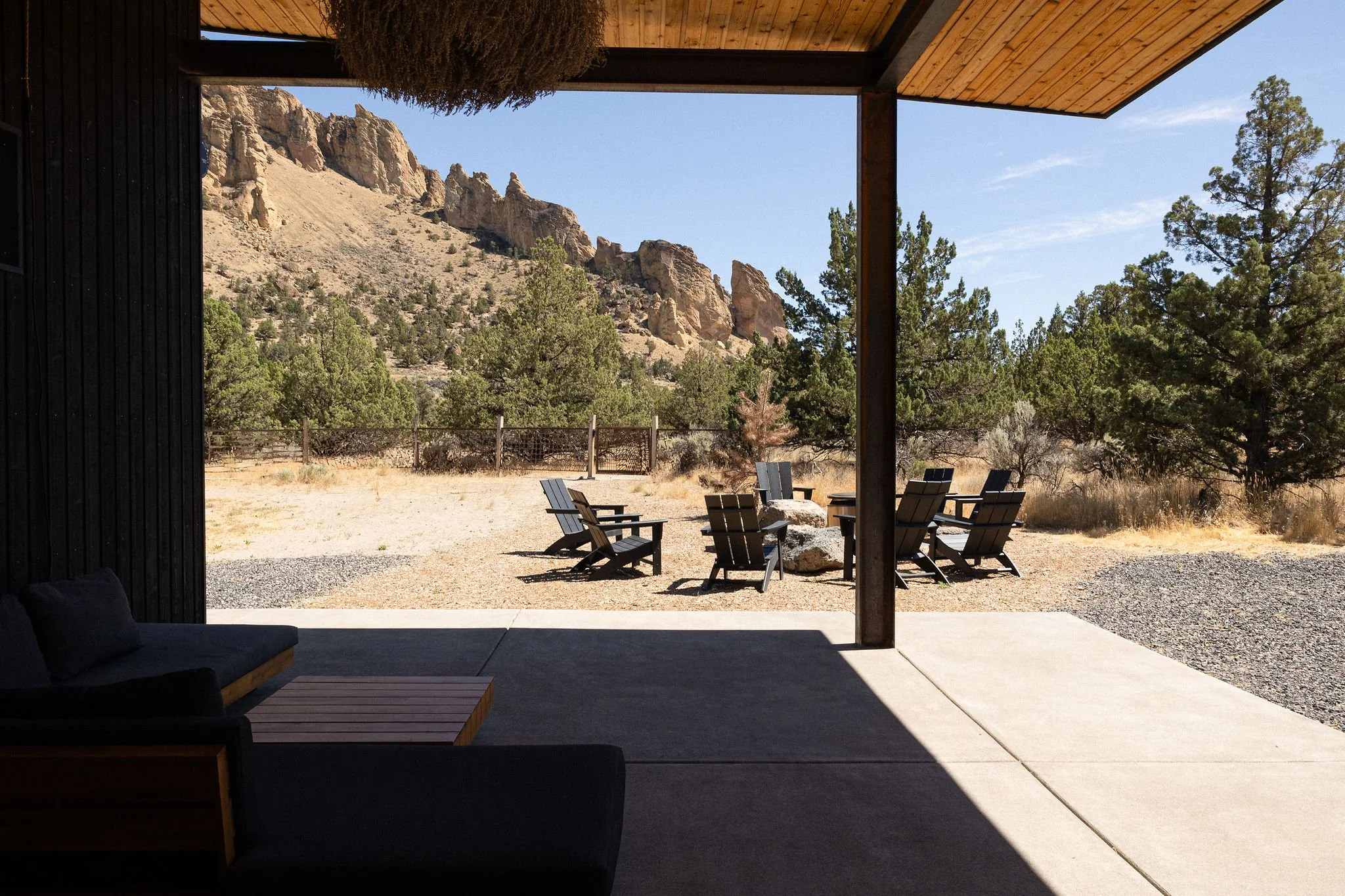 View from a covered patio looking out over a desert landscape with trees, rocks, and mountains in the background. There are several wooden chairs arranged around a fire pit outside, and a couch with cushions inside.