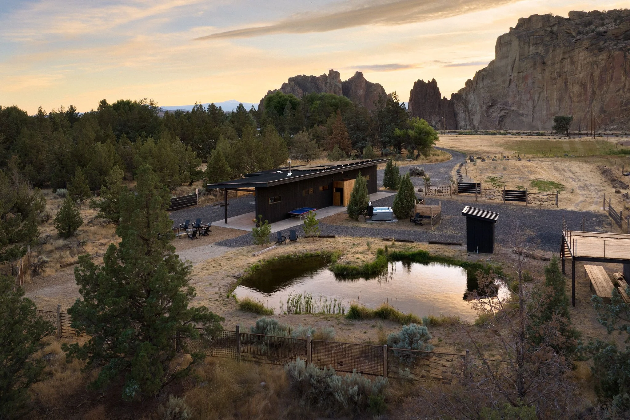 A rural landscape featuring a modern black house with outdoor seating, a small pond, and surrounding trees and mountains in the background during sunset.