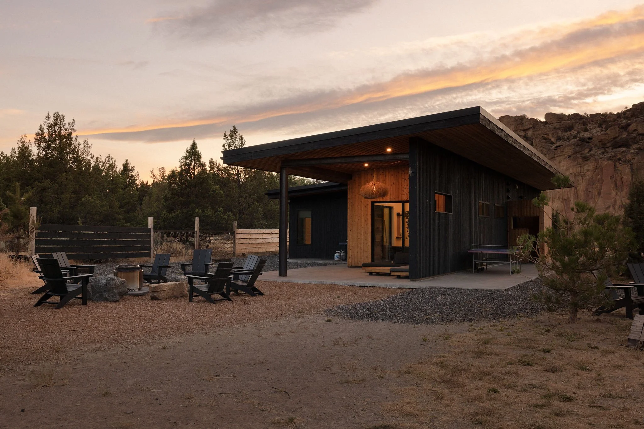 A modern house with black and wood exterior panels, large covered porch, surrounded by outdoor seating, situated in a desert landscape at sunset.