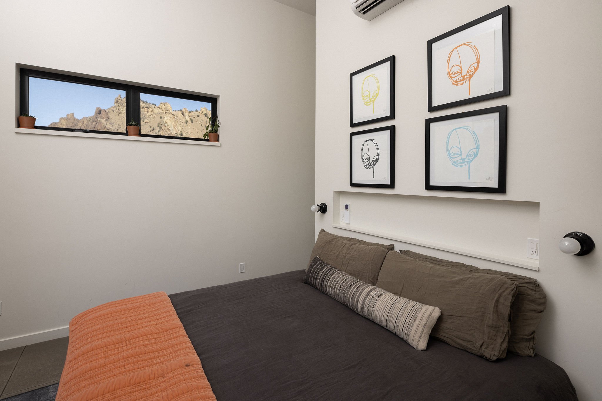 Bedroom with white walls, a horizontal window with three small potted plants, four framed colorful line art portraits of faces, a bed with brown bedding, one large pillow and a long striped bolster pillow, a wall-mounted air conditioning unit, and tw