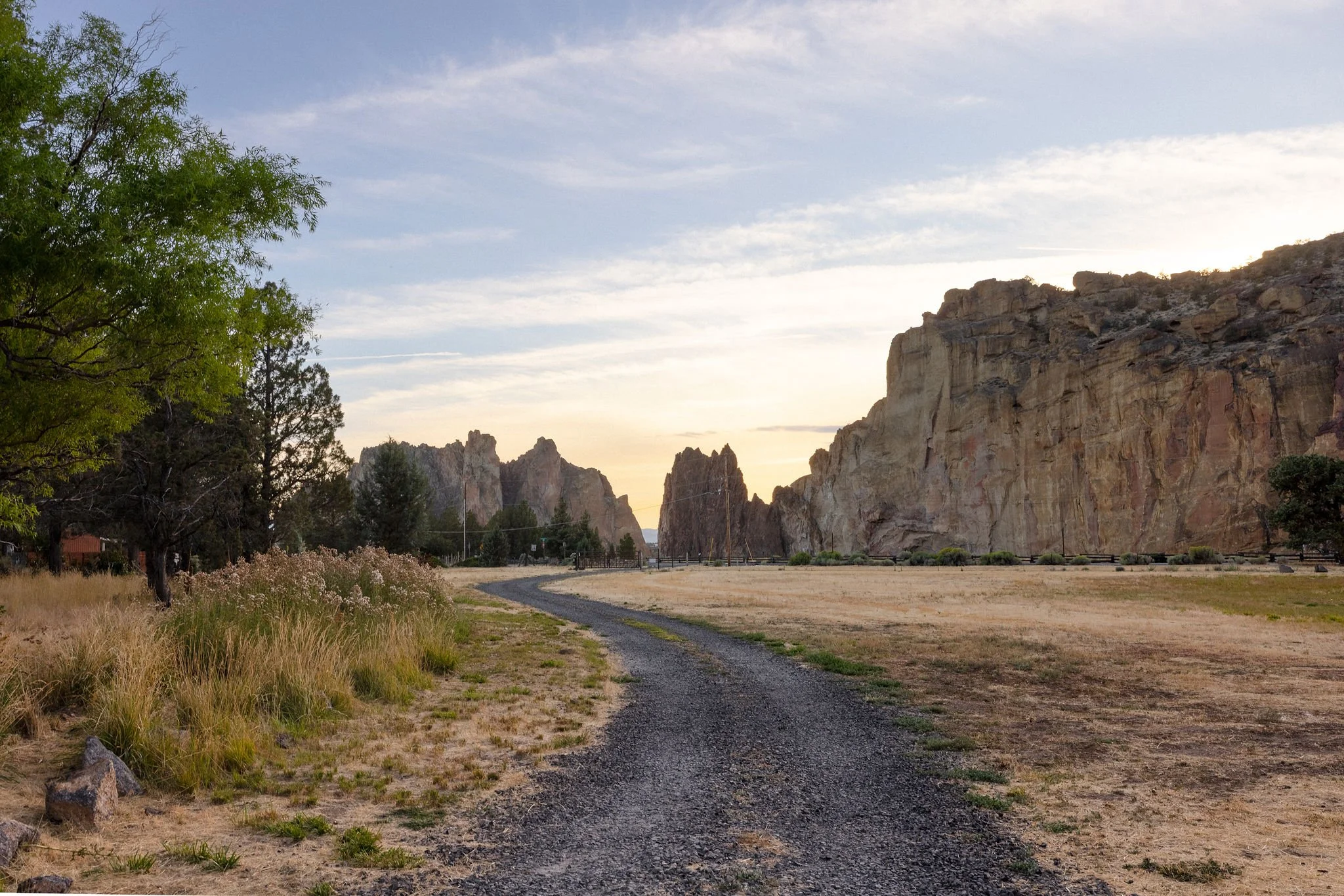 A dirt and gravel pathway winding through a semi-arid landscape with tall, rugged rock formations on the right and a few trees and grasses on the left, under a partly cloudy sky at sunset.