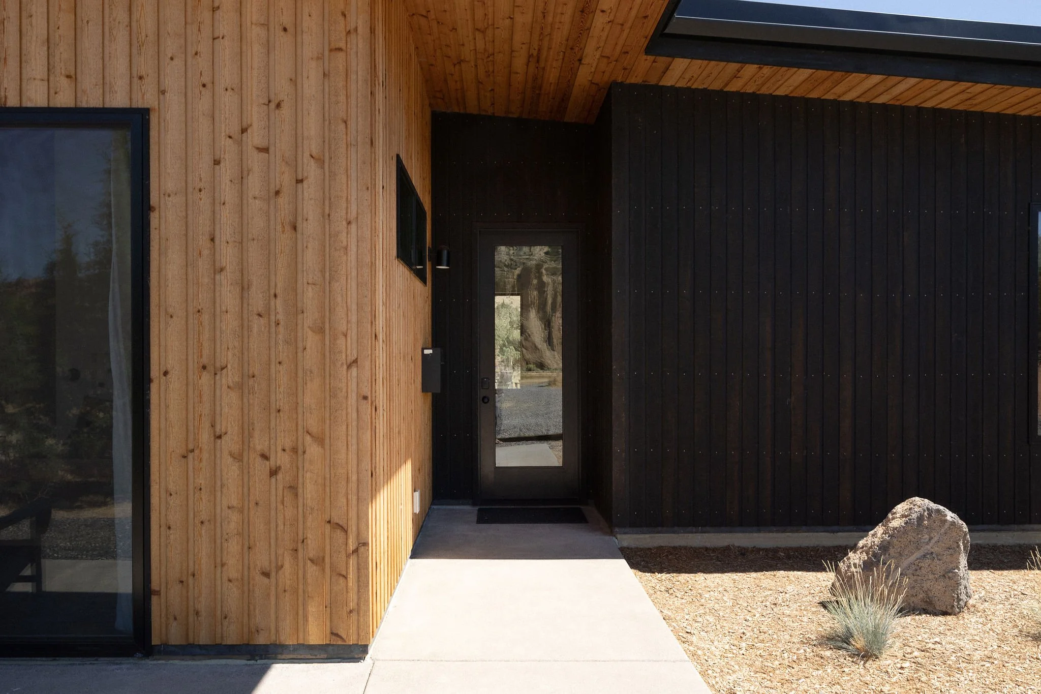 Modern house entrance with a black door, wooden siding on the left, dark vertical siding on the right, a concrete pathway, and a rock with desert plants outside.