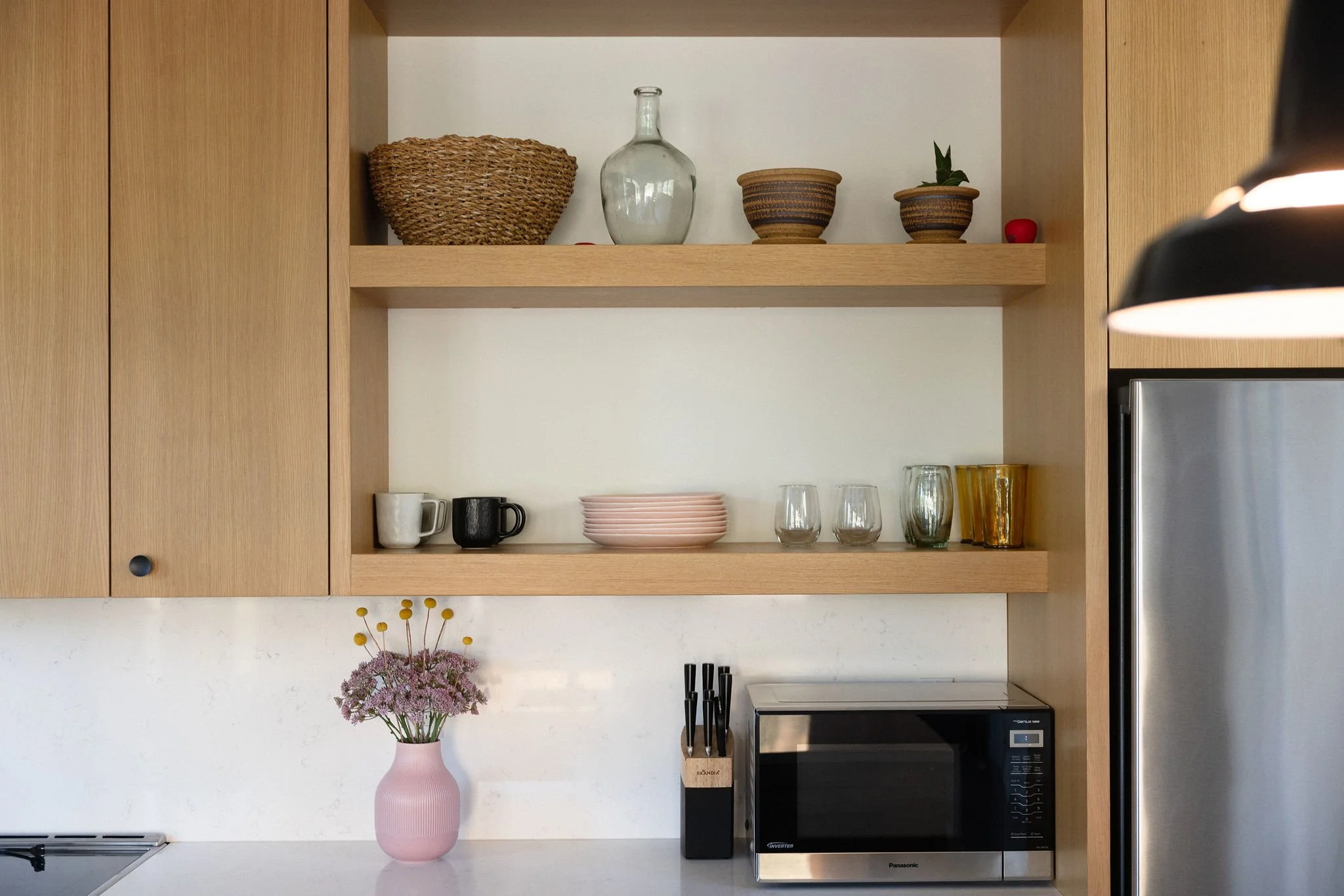 Open kitchen cabinet shelves with decorative items, cups, plates, and glasses, microwave on the counter, and a pink vase with flowers.