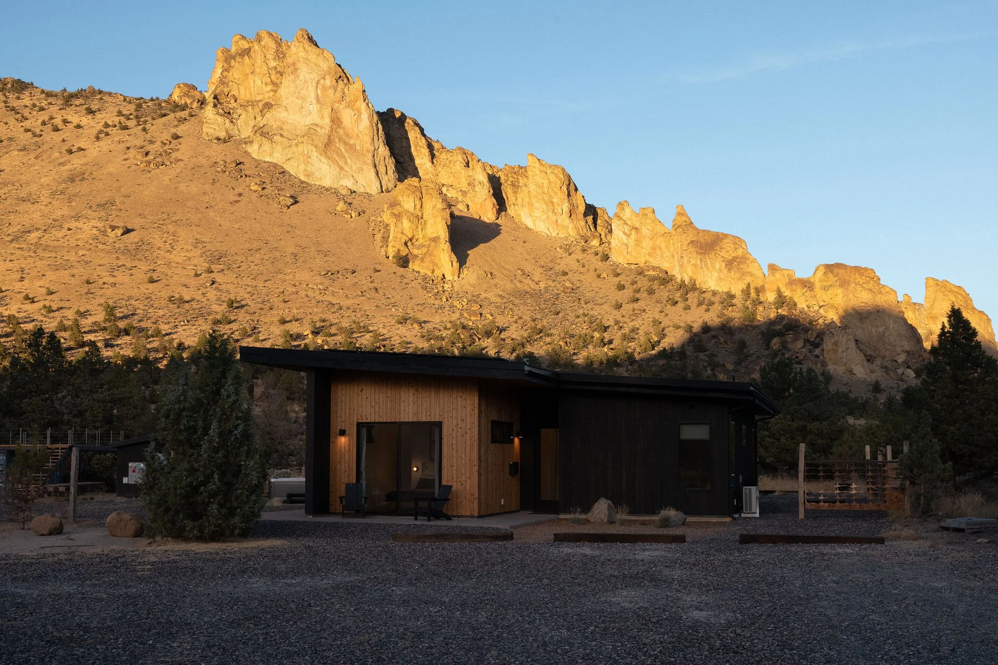 A modern house at sunset with mountains in the background, featuring black and wooden exterior walls, surrounded by desert landscape and sparse vegetation.