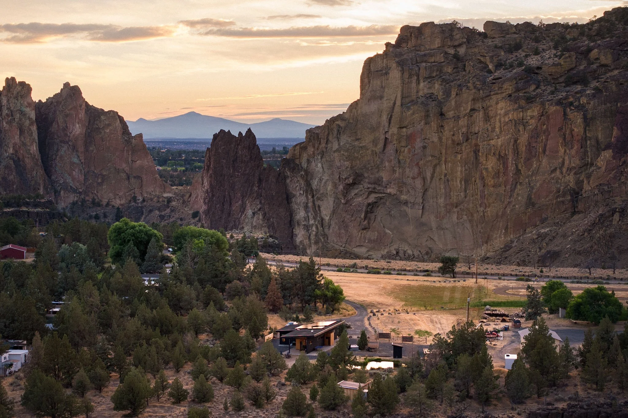 Mountain landscape at sunset with large rocky cliffs, trees, and a small residential area with several houses and parked cars in the foreground.