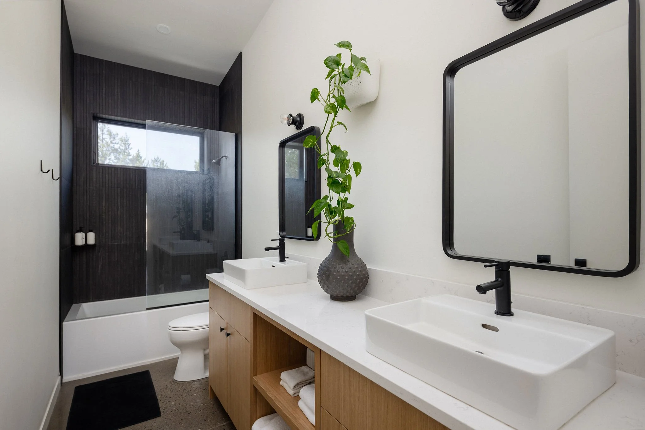 Modern bathroom with white double sinks, black faucets, large mirrors, a wooden vanity with open shelves and towels, a decorative vase with a green plant, a toilet, and a glass-enclosed shower area with a window