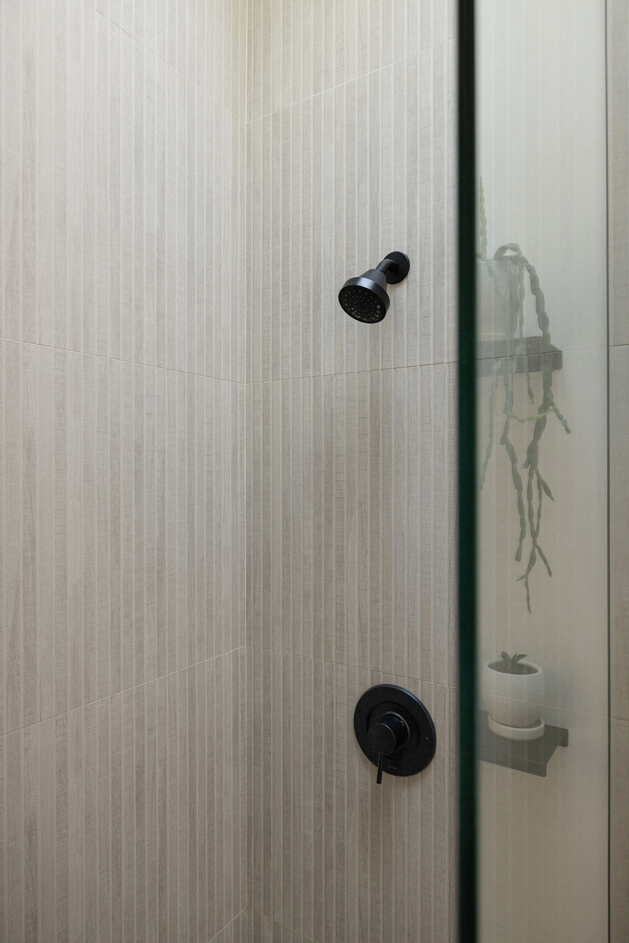 A minimalist bathroom shower with beige tiled walls, a black showerhead, and a black control knob. Reflection shows a hanging plant in a white pot.
