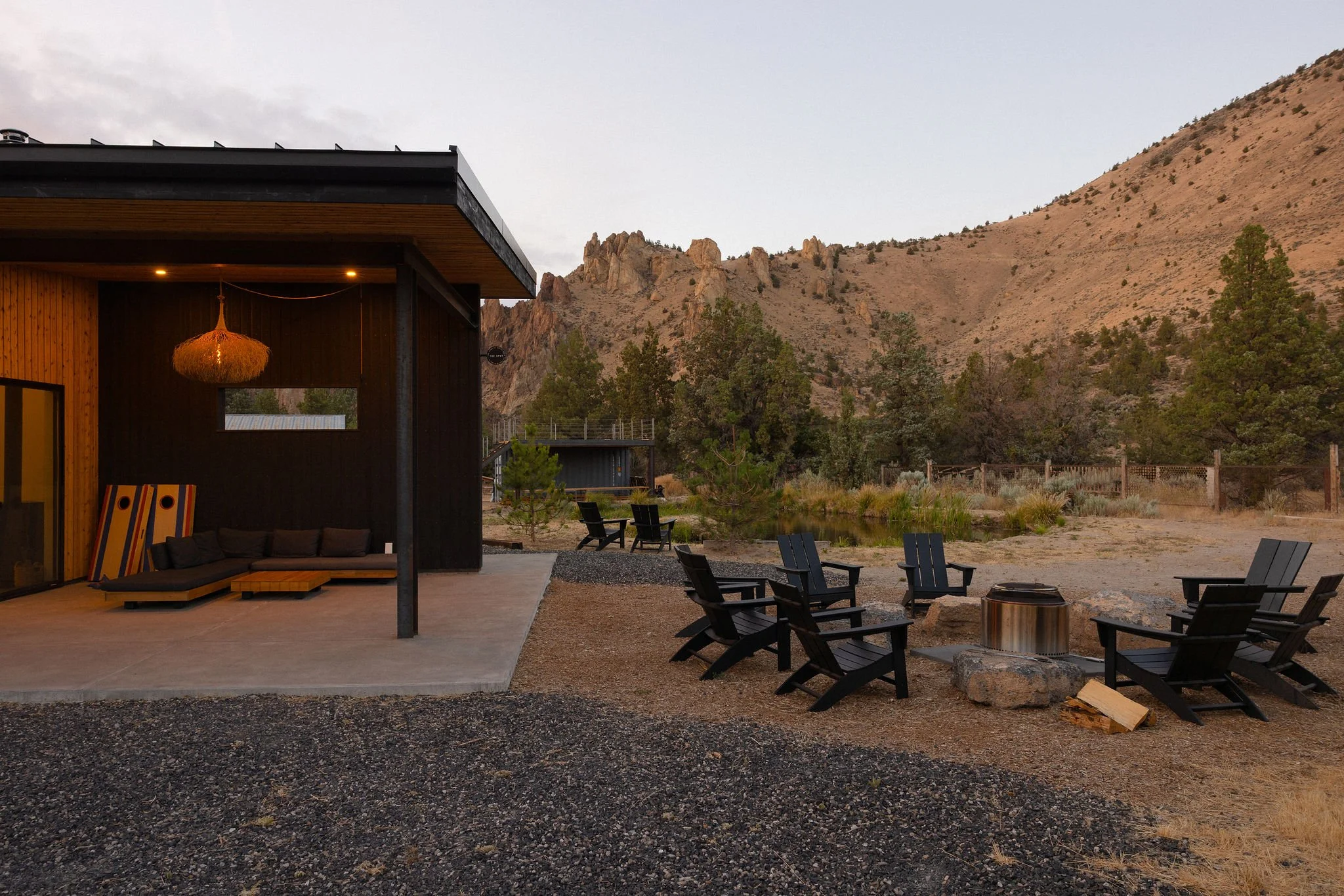 Outdoor patio area with black Adirondack chairs arranged around a fire pit, a covered porch with a hanging wicker light, and a mountain landscape in the background with trees and a fence.