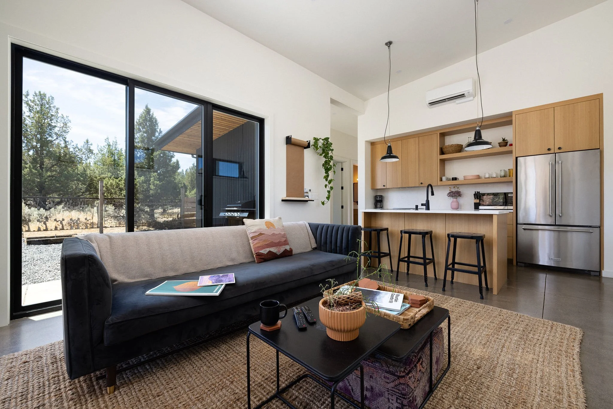 Living room with large sliding glass door, black sofa with beige and patterned pillows, wooden coffee table with plant, mugs, TV remote, and magazines, open kitchen with wooden cabinets, black pendant lights, and stainless steel refrigerator, natural