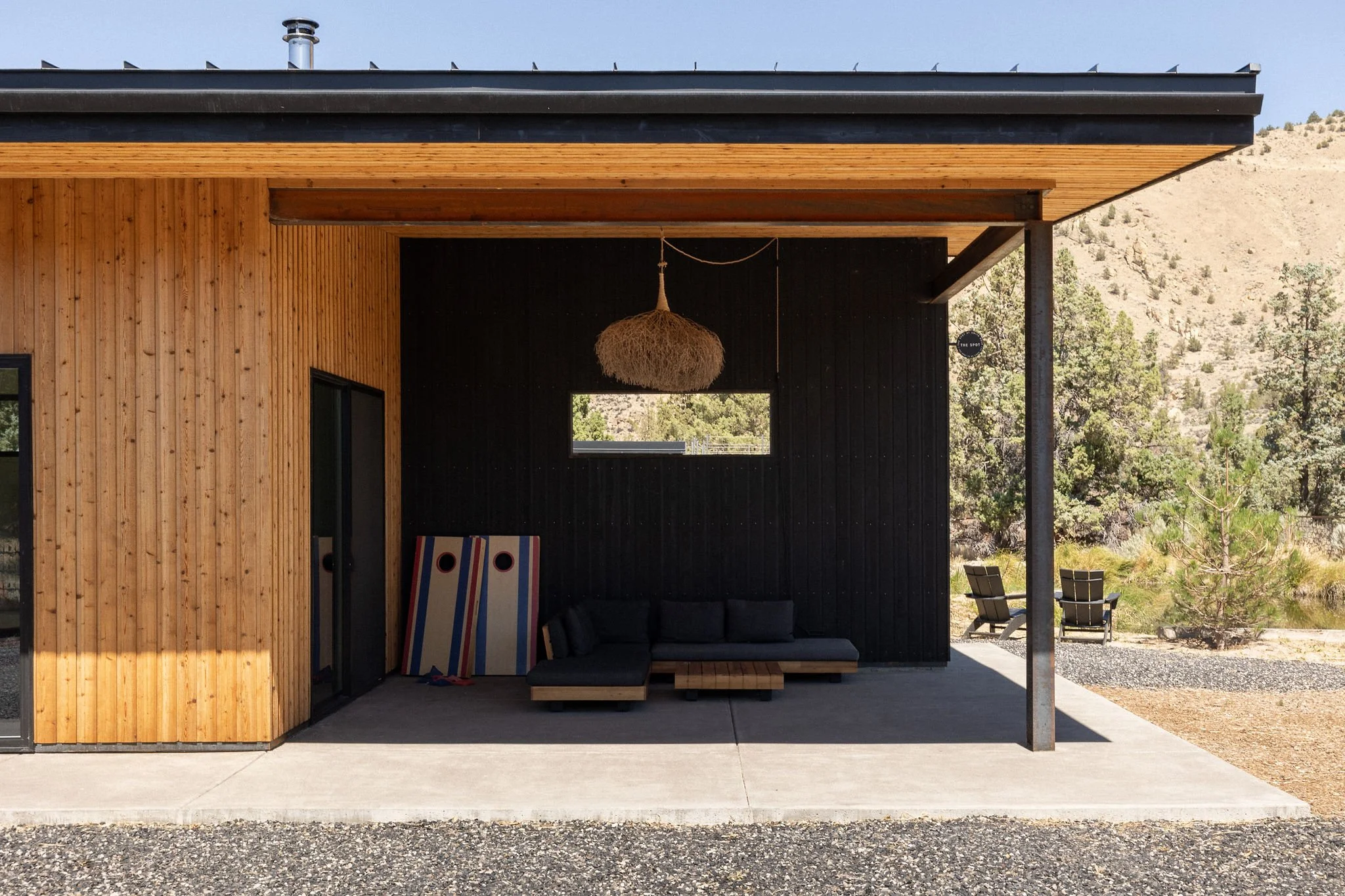 Exterior view of a modern house with a covered patio, wooden and black wall design, cushioned seating, hanging light fixture, and outdoor chairs in a natural landscape.