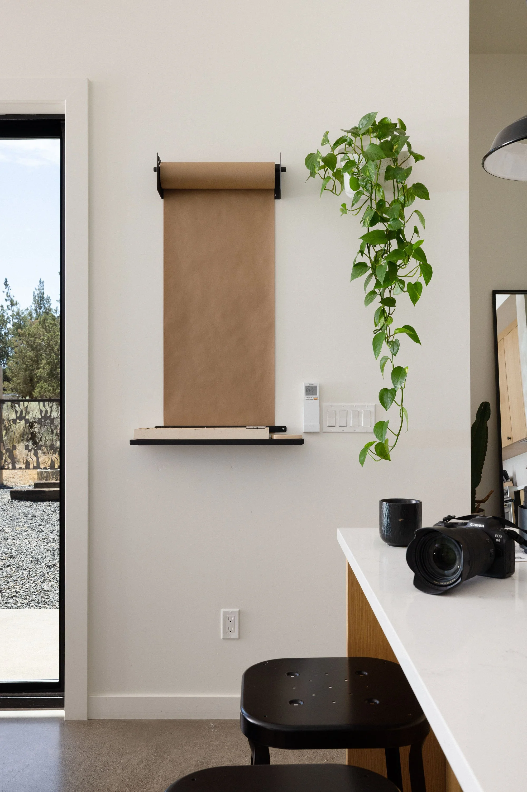 Interior view of a modern kitchen or dining area with a white countertop, black stool, camera, black cup, green hanging plant, and a section of outdoor patio visible through a glass door.