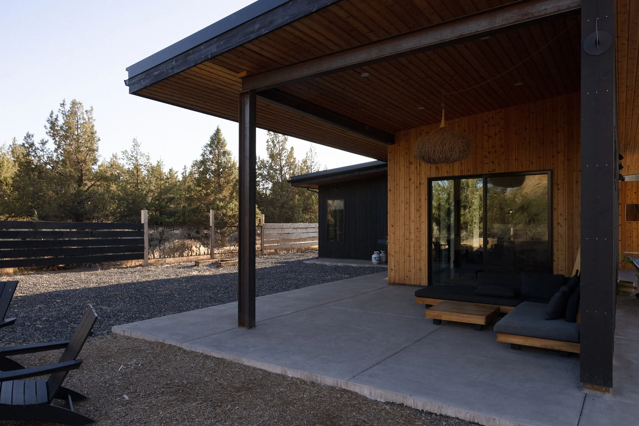 Modern house with a covered patio, black and wood exterior, outdoor seating, and trees in the background.