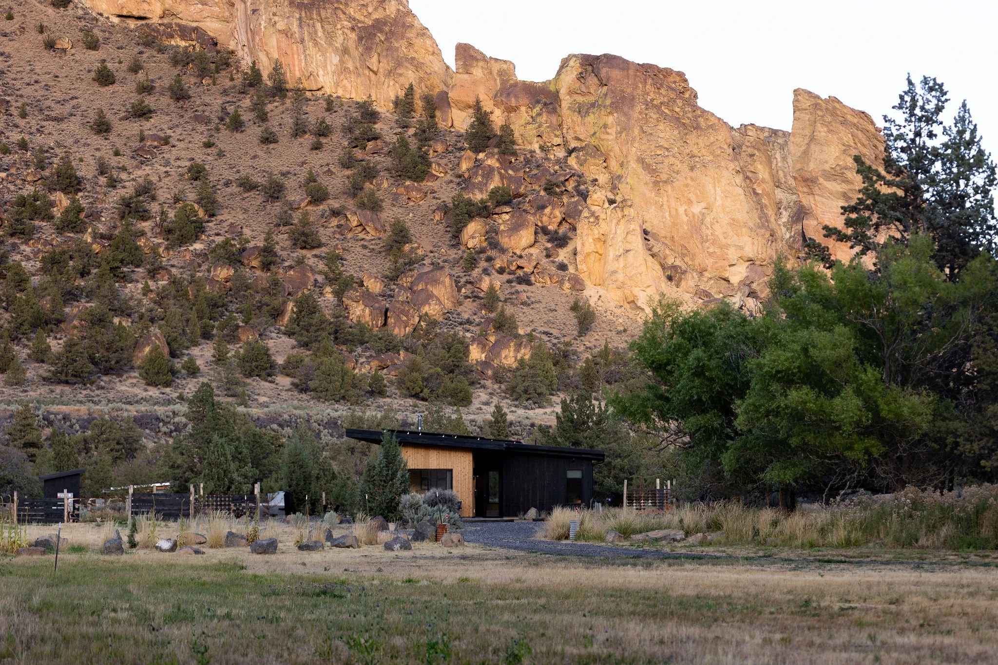 A modern black and wood house situated at the base of a rocky mountain with sparse trees and grass in the foreground.