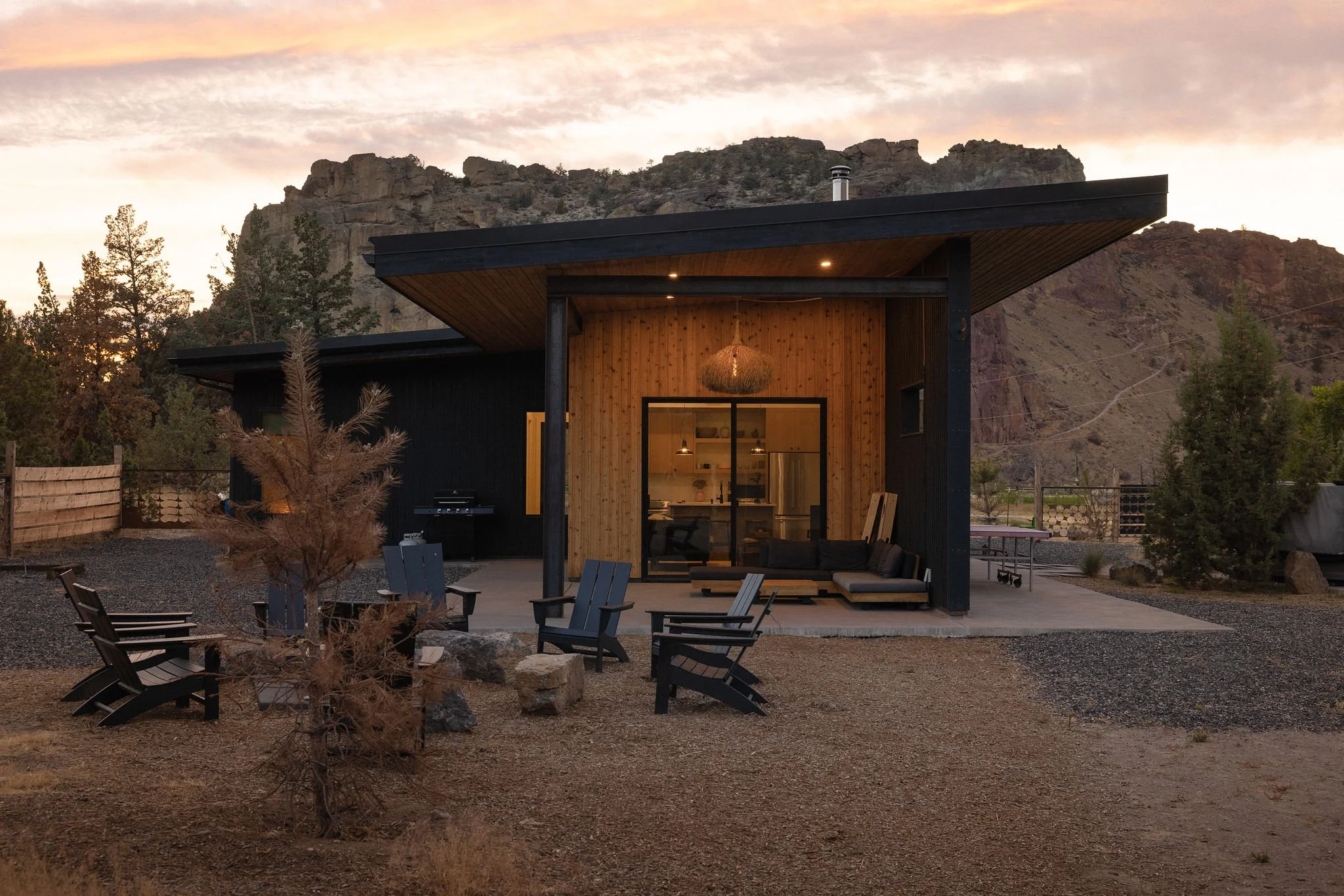 Modern house with an outdoor patio, surrounded by trees and mountains at sunset, featuring seating, a small tree with brown leaves, and a black and wood exterior.