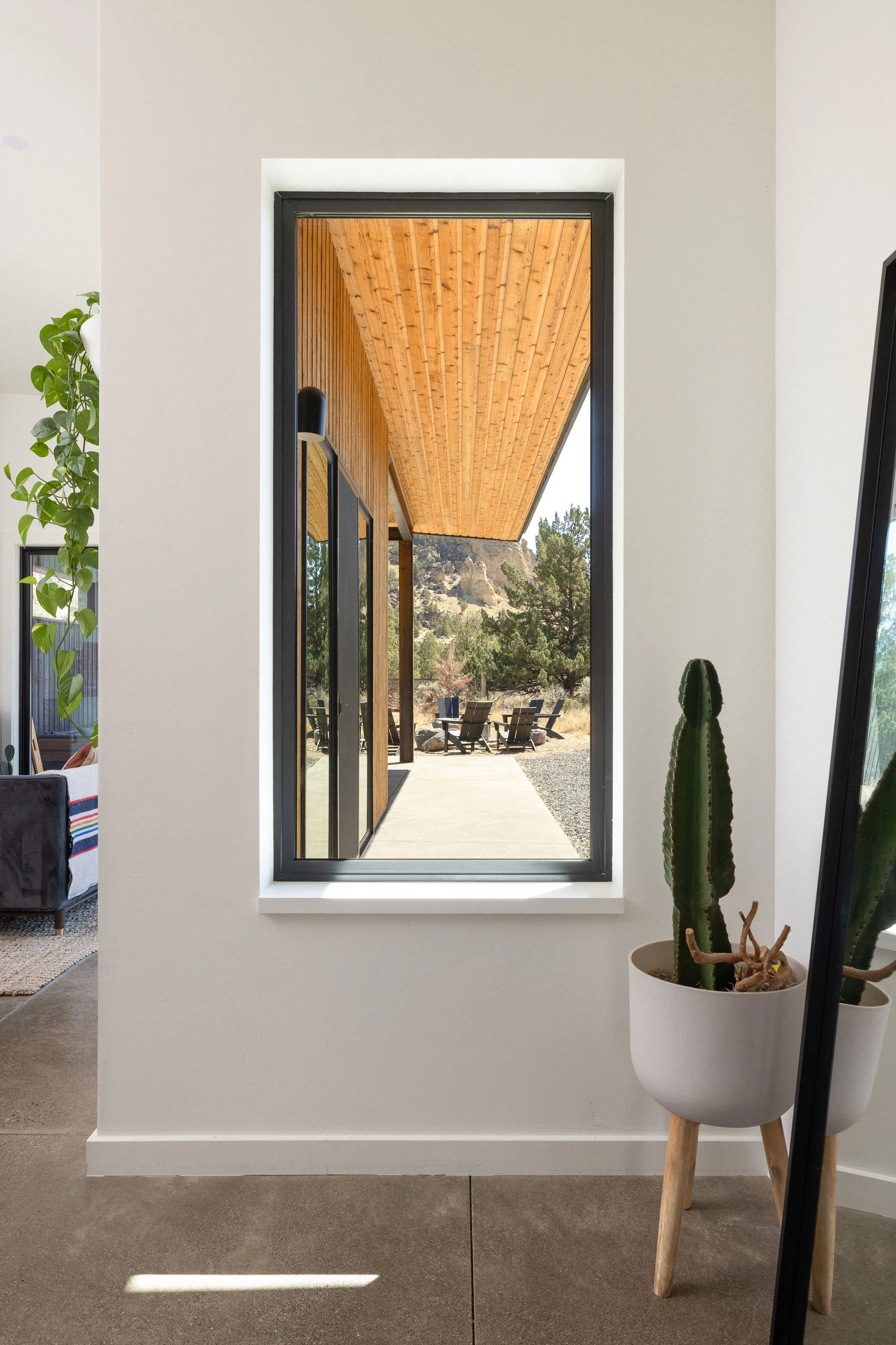 A view through a rectangular window showing a covered outdoor patio with wooden ceiling, trees, and outdoor chairs.