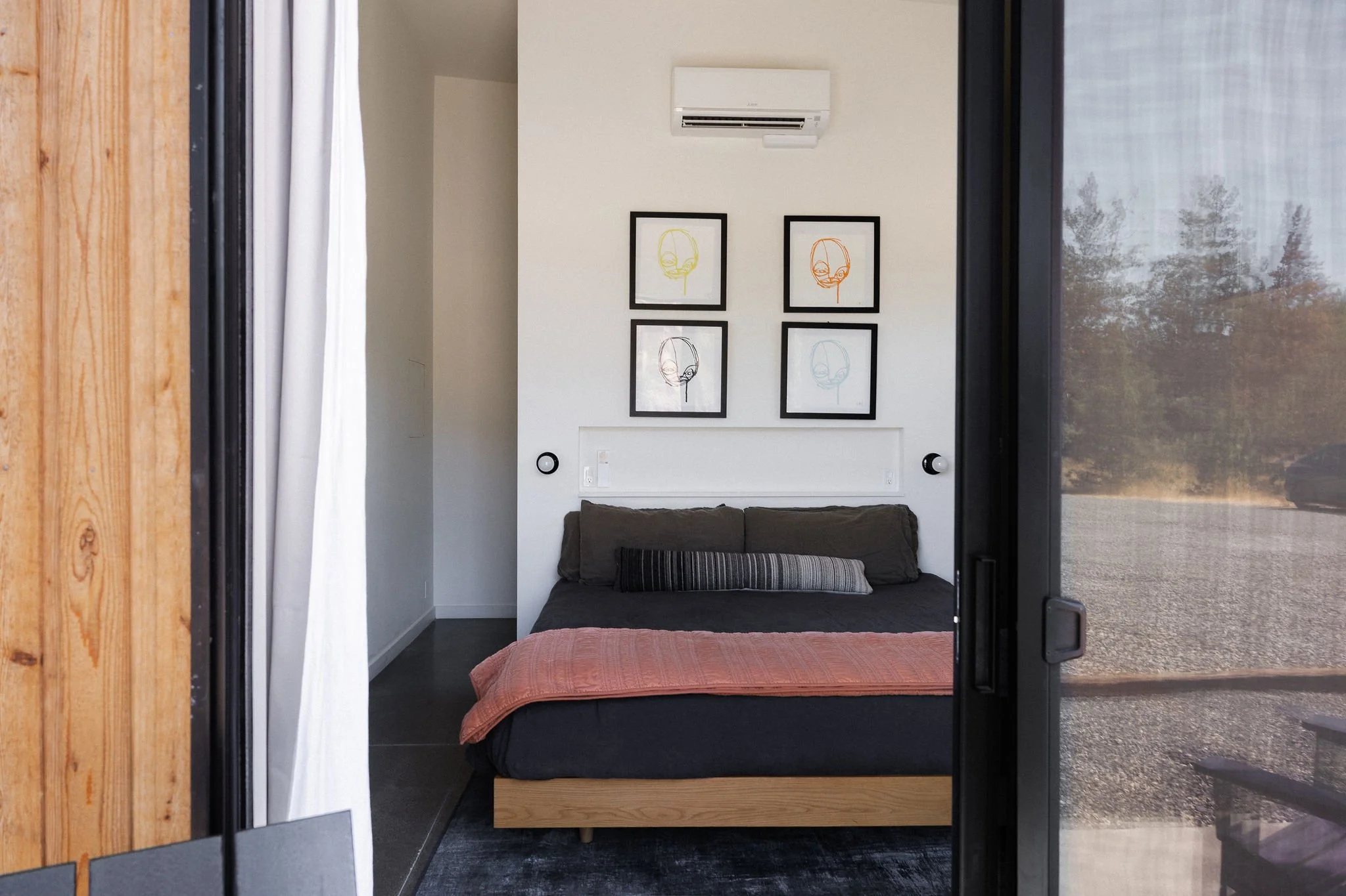 Modern bedroom with black bed frame, dark bedding, and orange throw, framed abstract art on white wall, and glass sliding door with outdoor view.