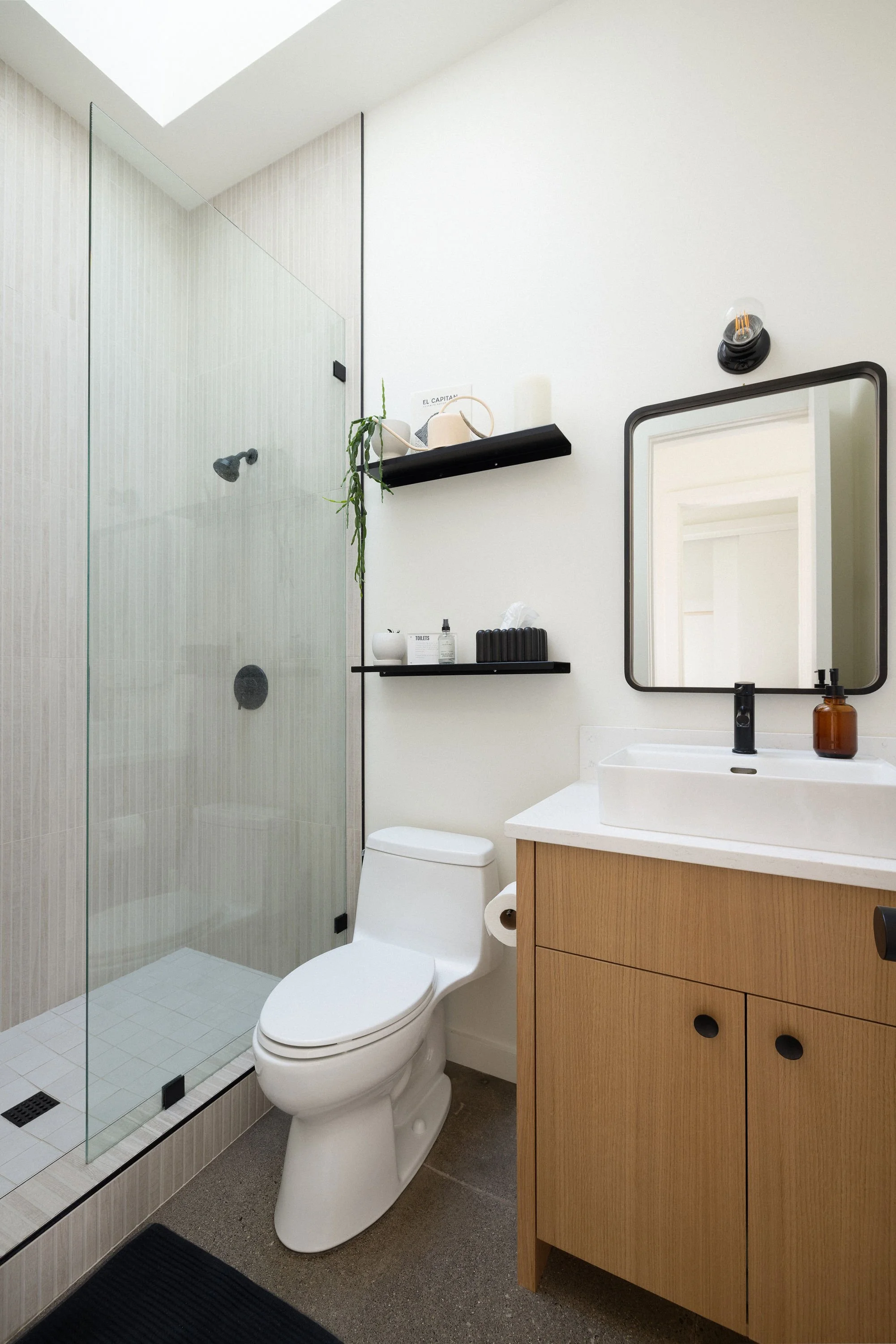 Modern bathroom with a glass shower, white toilet, wooden vanity with white sink, mirror, black fixtures, and black shelving with decor.