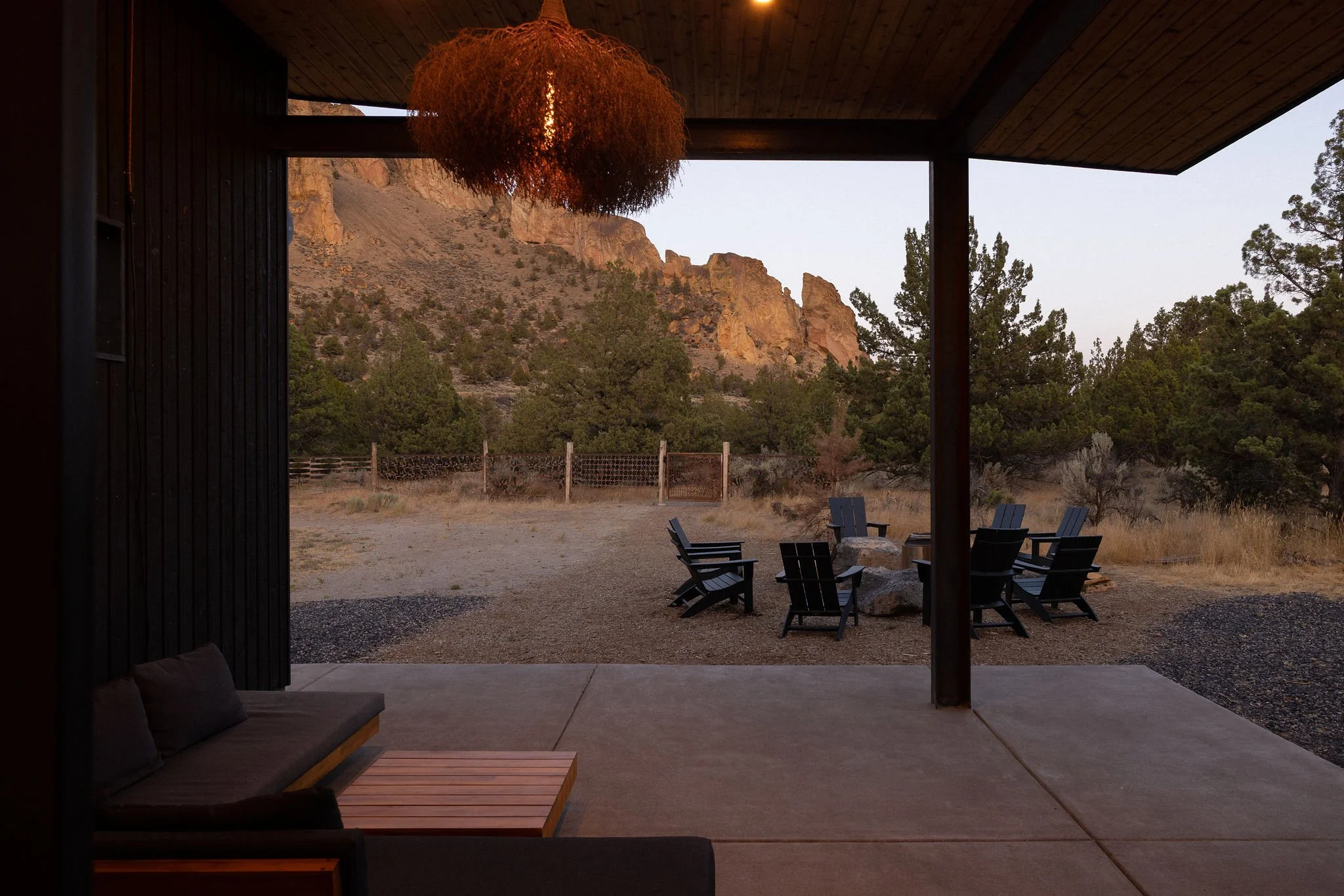 A covered patio with outdoor seating overlooking a rocky, mountainous landscape with trees and a clear sky at sunset.