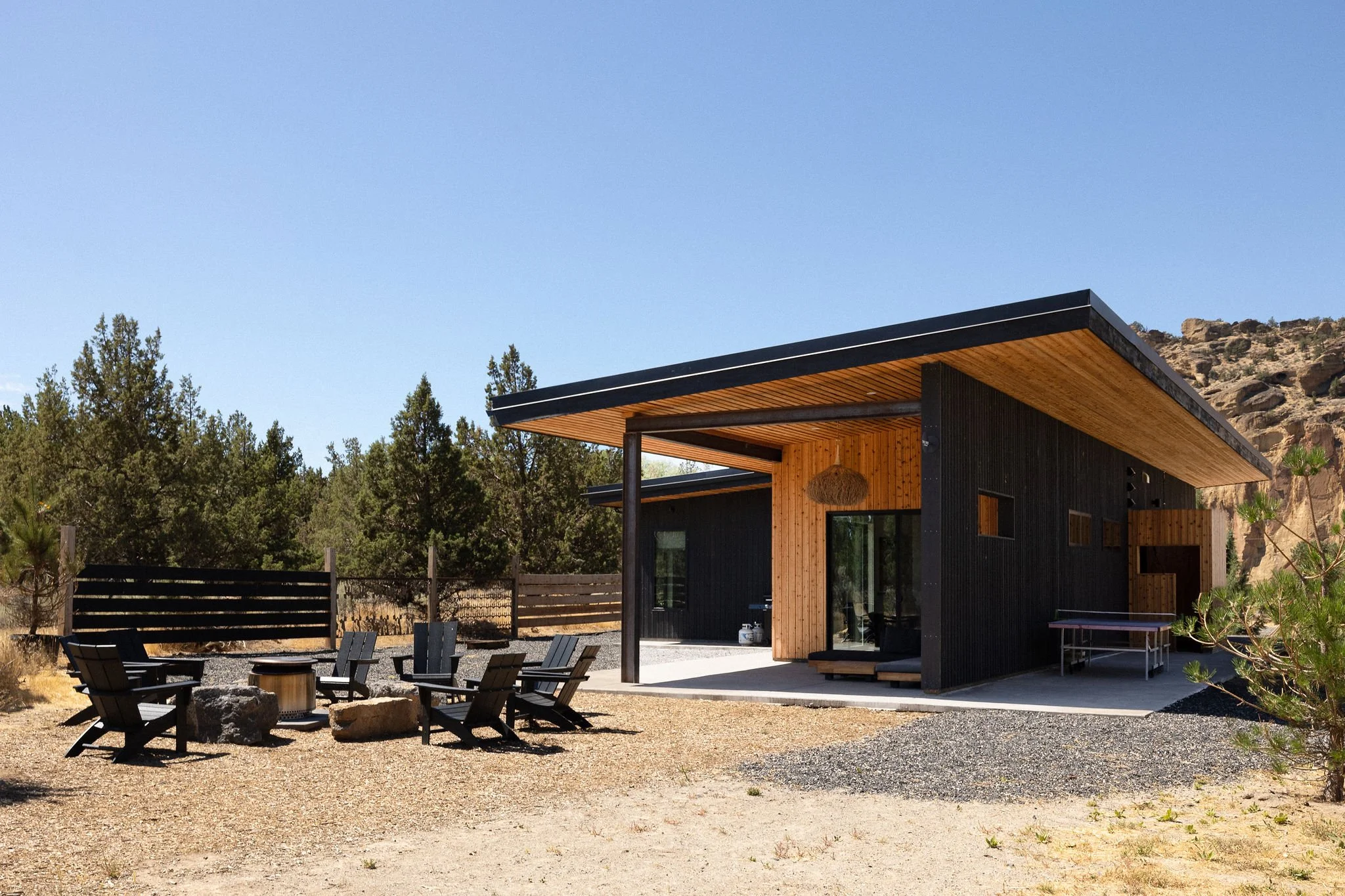 Modern house with black and wood exterior, outdoor seating with Adirondack chairs and a table, in a desert or dry landscape with trees and rocky hills in the background.
