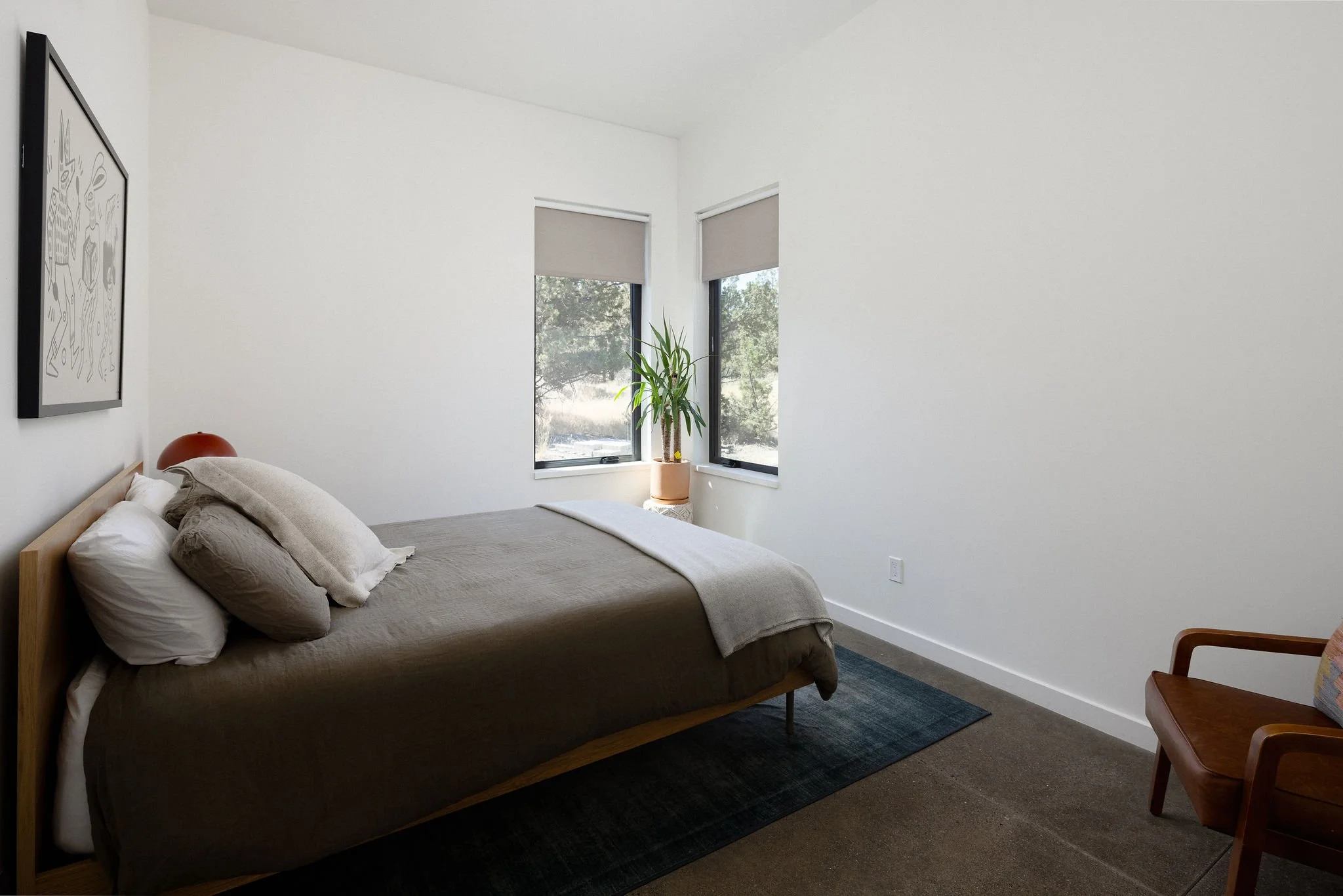 Minimalist bedroom with a bed, a plant by the window, a chair, and wall art, featuring neutral tones and natural light.