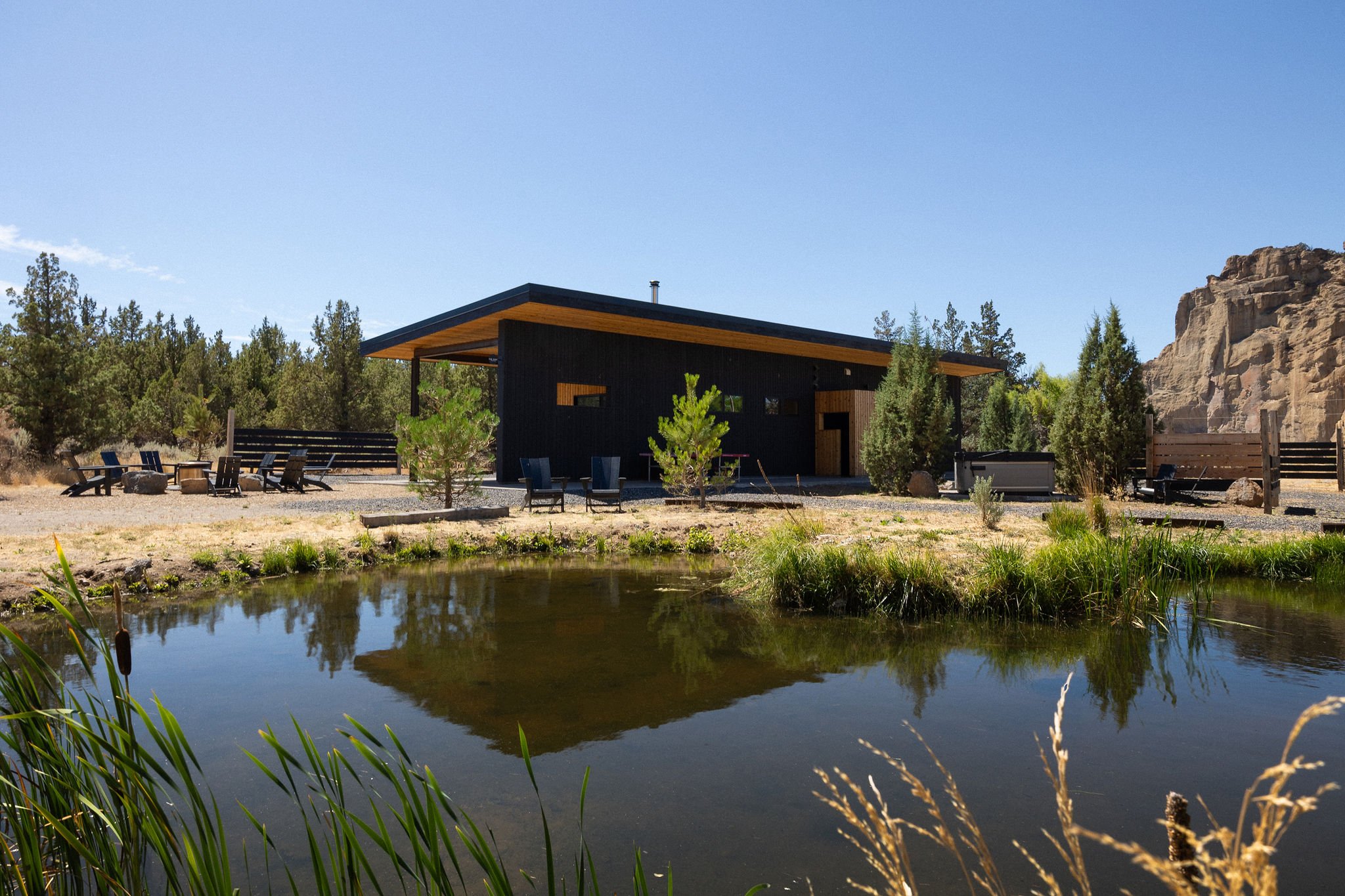 Modern black house with a flat roof surrounded by trees, with a small pond in the foreground reflecting the house, under a clear blue sky.
