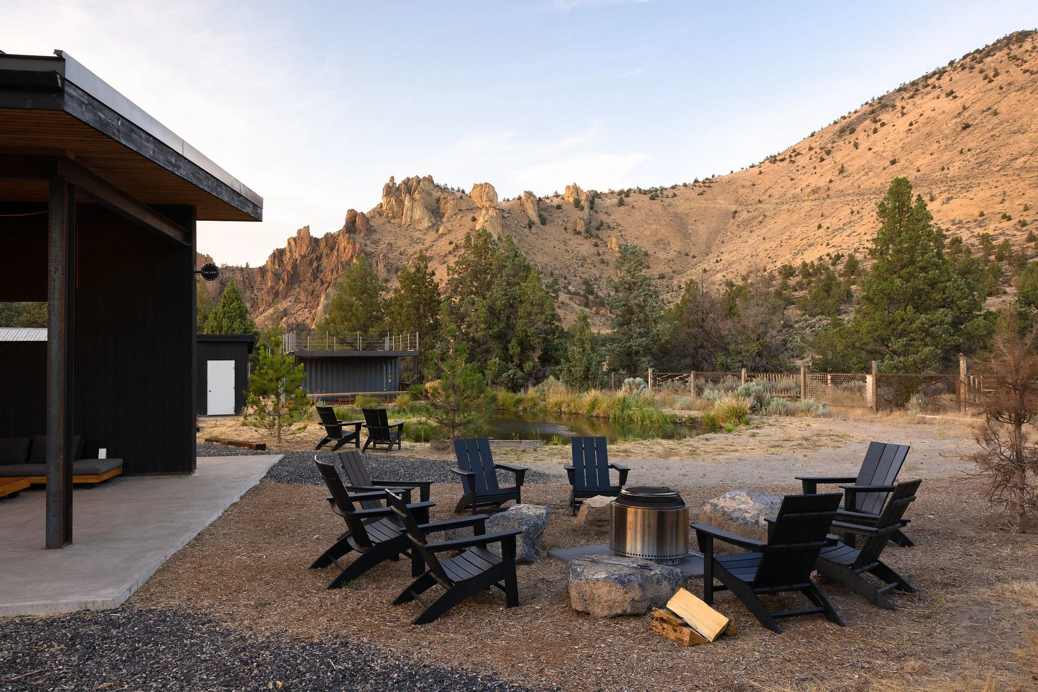 Outdoor patio with black Adirondack chairs arranged in a circle around a fire pit, set against a mountainous landscape with trees and a small pond.